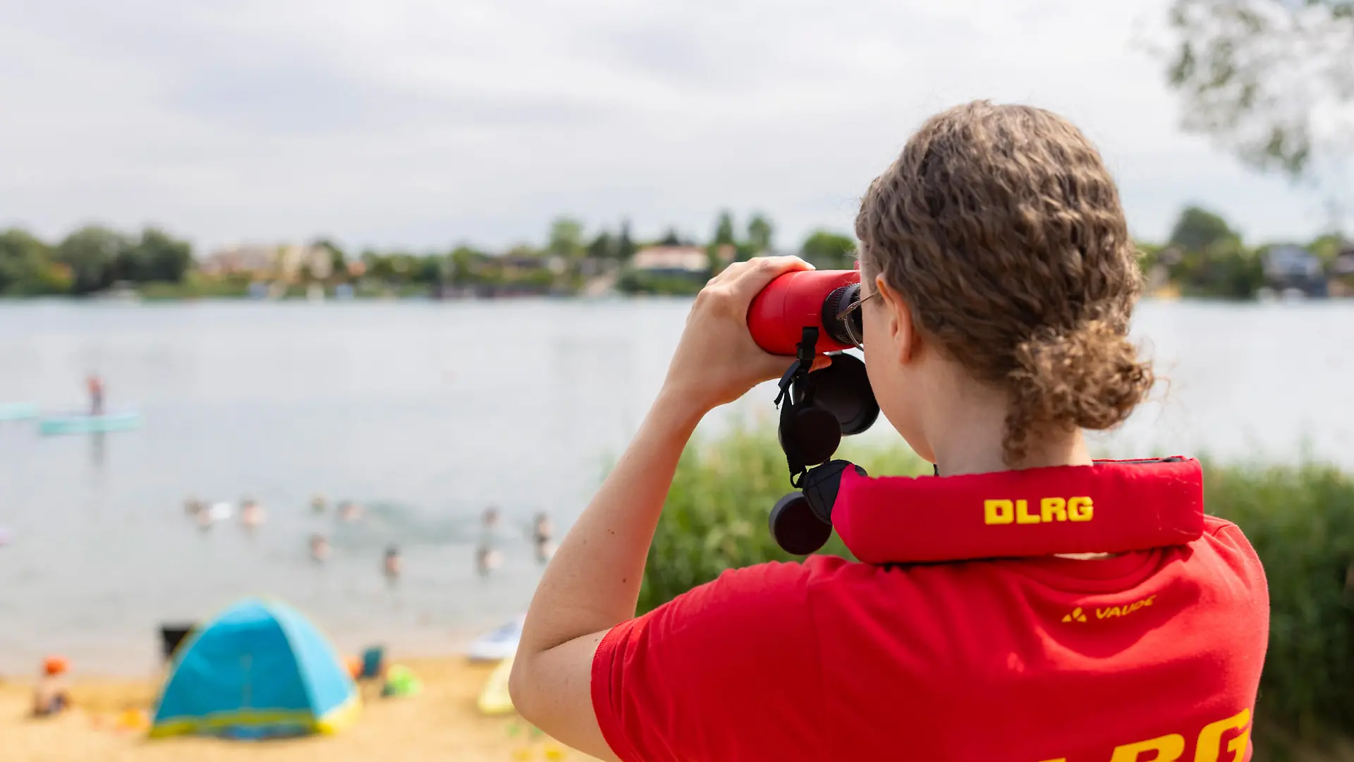 Rettungsschwimmerin der DLRG-Wasserrettung beobachtet durch ein Fernglas die Badegaeste am Alperstedter See in Thueringen (Foto vom 05.07.2025). Die Wachstation am Alperstedter See bei Erfurt wird durch Rettungsschwimmer aus dem DLRG Landesverband Thueringen besetzt. Die Saison laeuft vom 29. Mai bis 14. September (je nach Wetterlage). Die DLRG Teams halten Wache, beobachten den Badesee aktiv und sorgen fuer die Sicherheit der Badegaeste. Im Notfall greifen sie ein  Personen, die in Gefahr geraten sind, werden schnellstmoeglich aus dem Wasser gerettet. Die Ausstattung umfasst dazu unter anderem Rettungsboje, Rettungsboard, Gurtretter sowie Wurfsack. Kleinere Verletzungen oder Medizin Notfaelle werden direkt vor Ort im SAN Bereich versorgt.