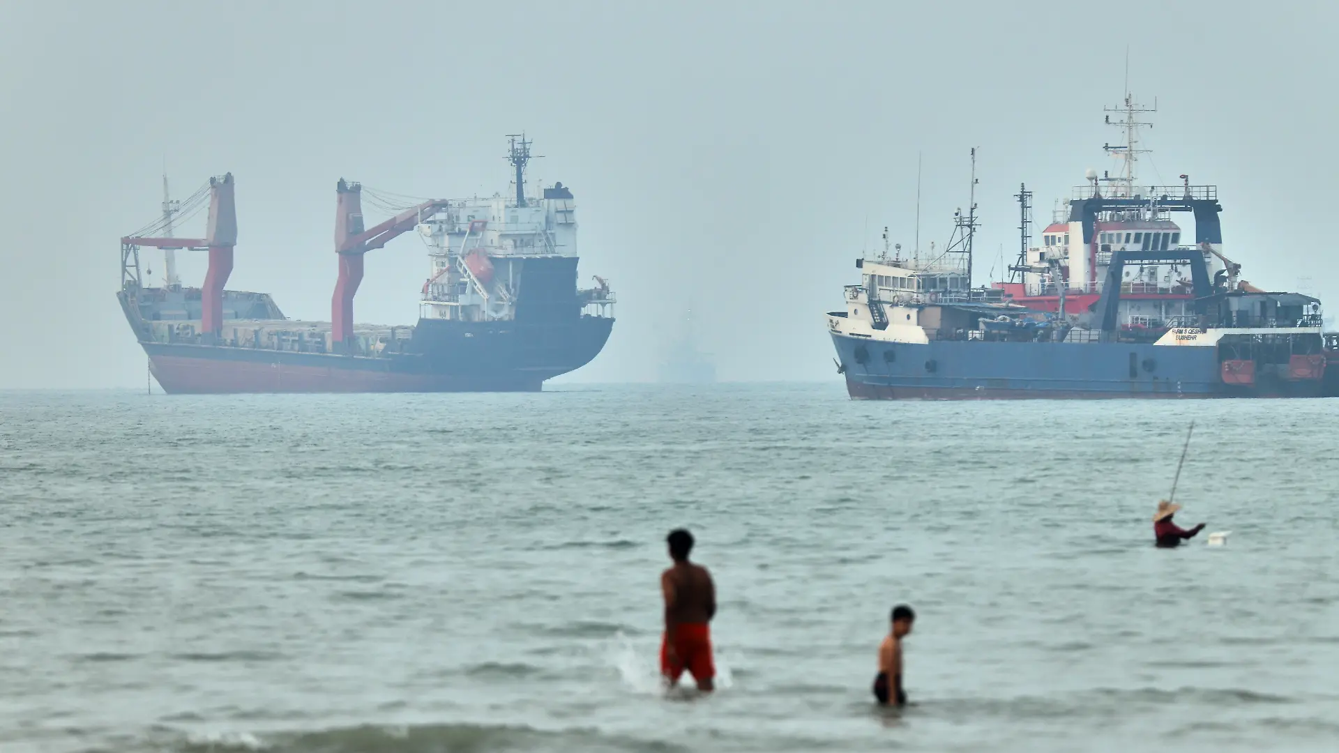 Kinder waten im Wasser. Im Hintergrund liegen vor Anker befindliche Frachtschiffe in der Straße von Hormus. (zu dpa: «Sorge vor US-Angriffen überschattet Staatsbegräbnis im Iran») +++ dpa-Bildfunk +++