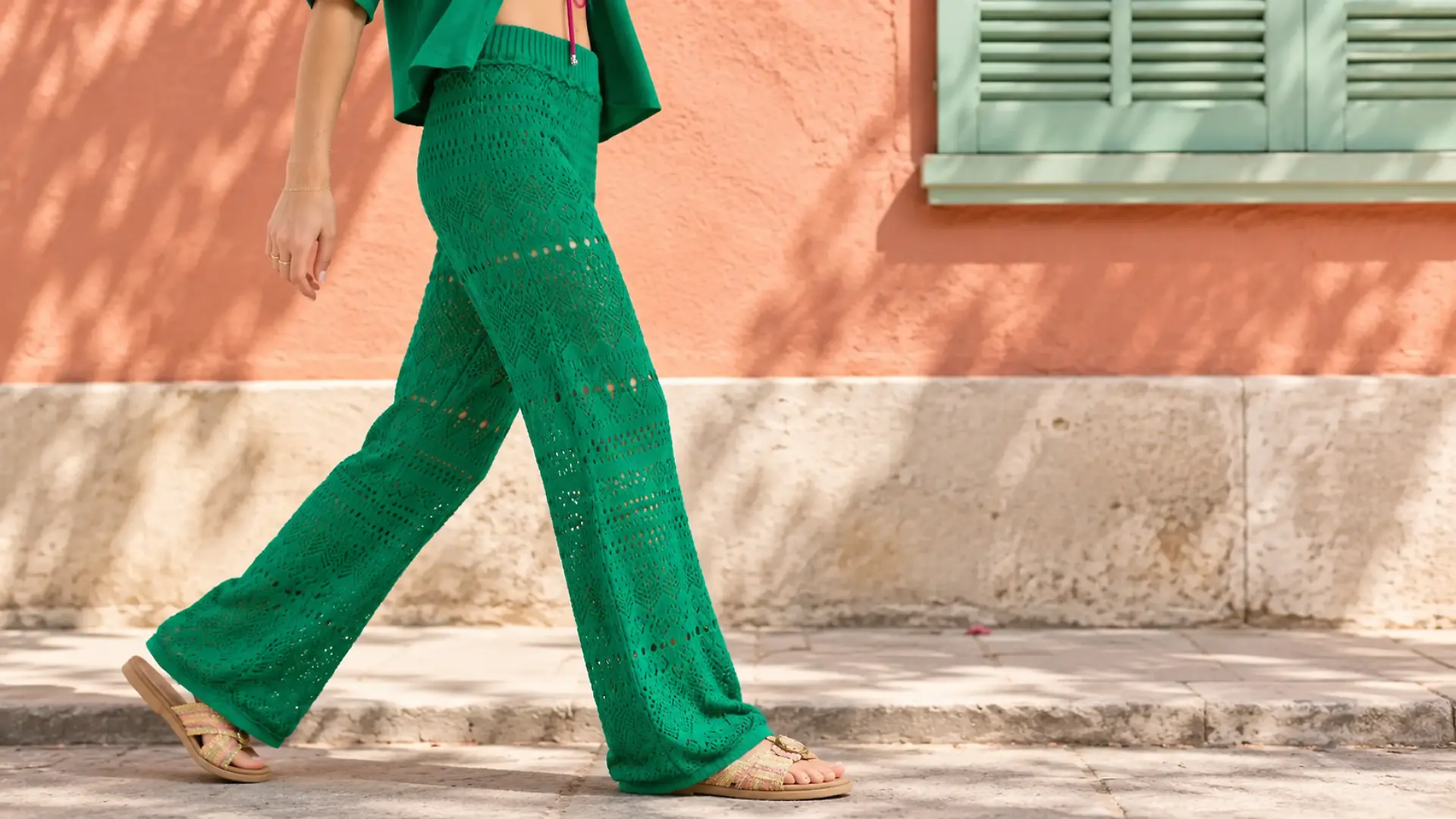 Ein Model trägt eine grüne Häkelhose und läuft vor einer farbigen Hausfassade entlang.