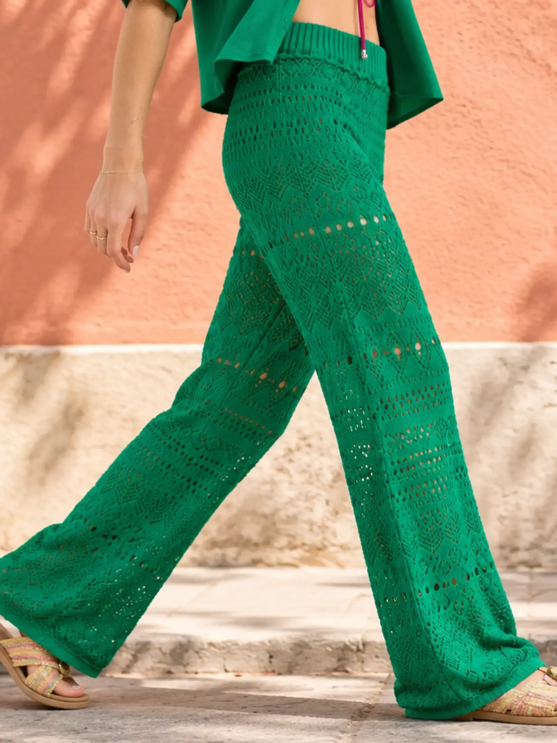 Ein Model trägt eine grüne Häkelhose und läuft vor einer farbigen Hausfassade entlang.