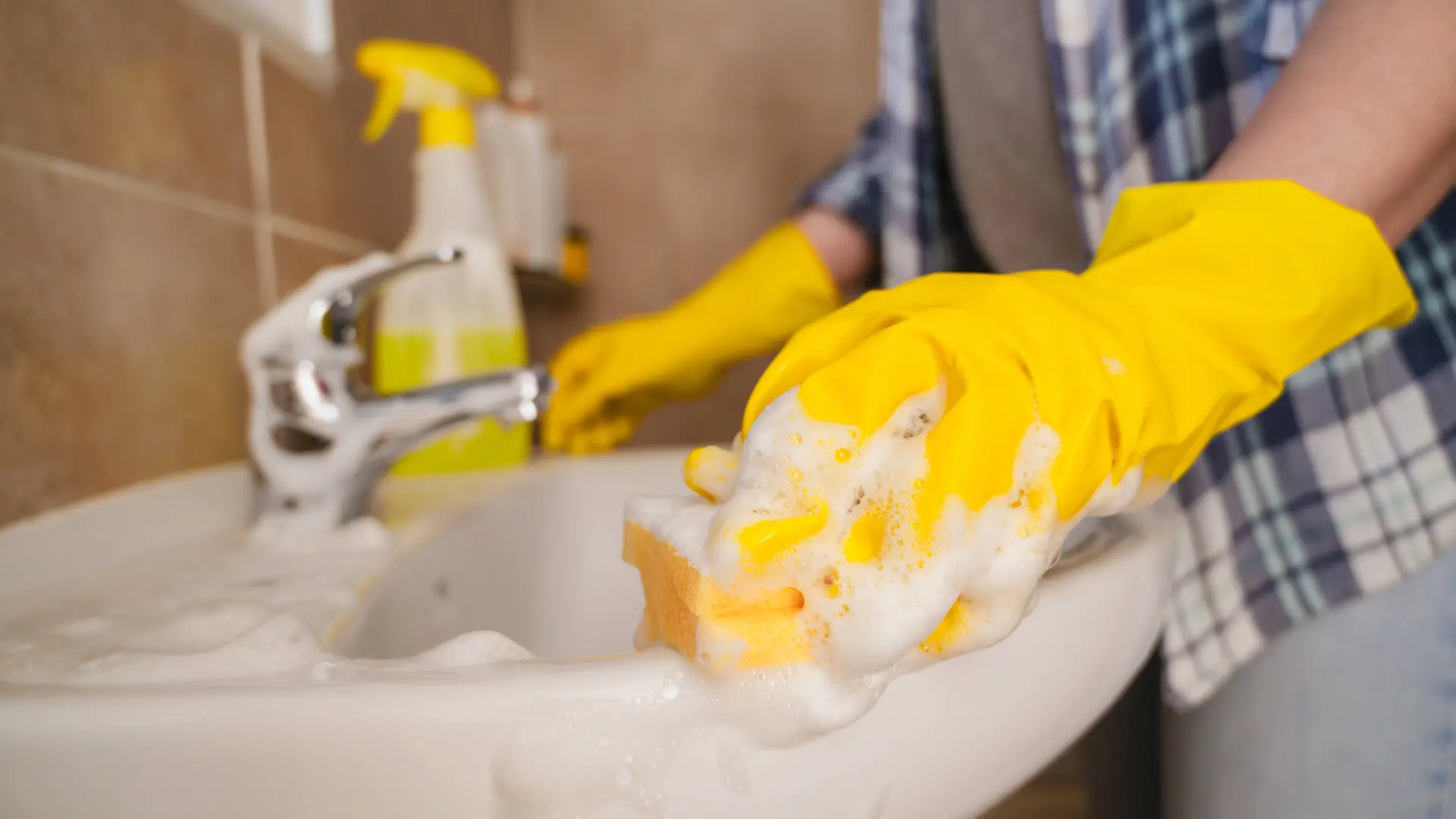Frau reinigt Waschbecken im Badezimmer mit Gummihandschuh und Schwamm – Symbolbild
