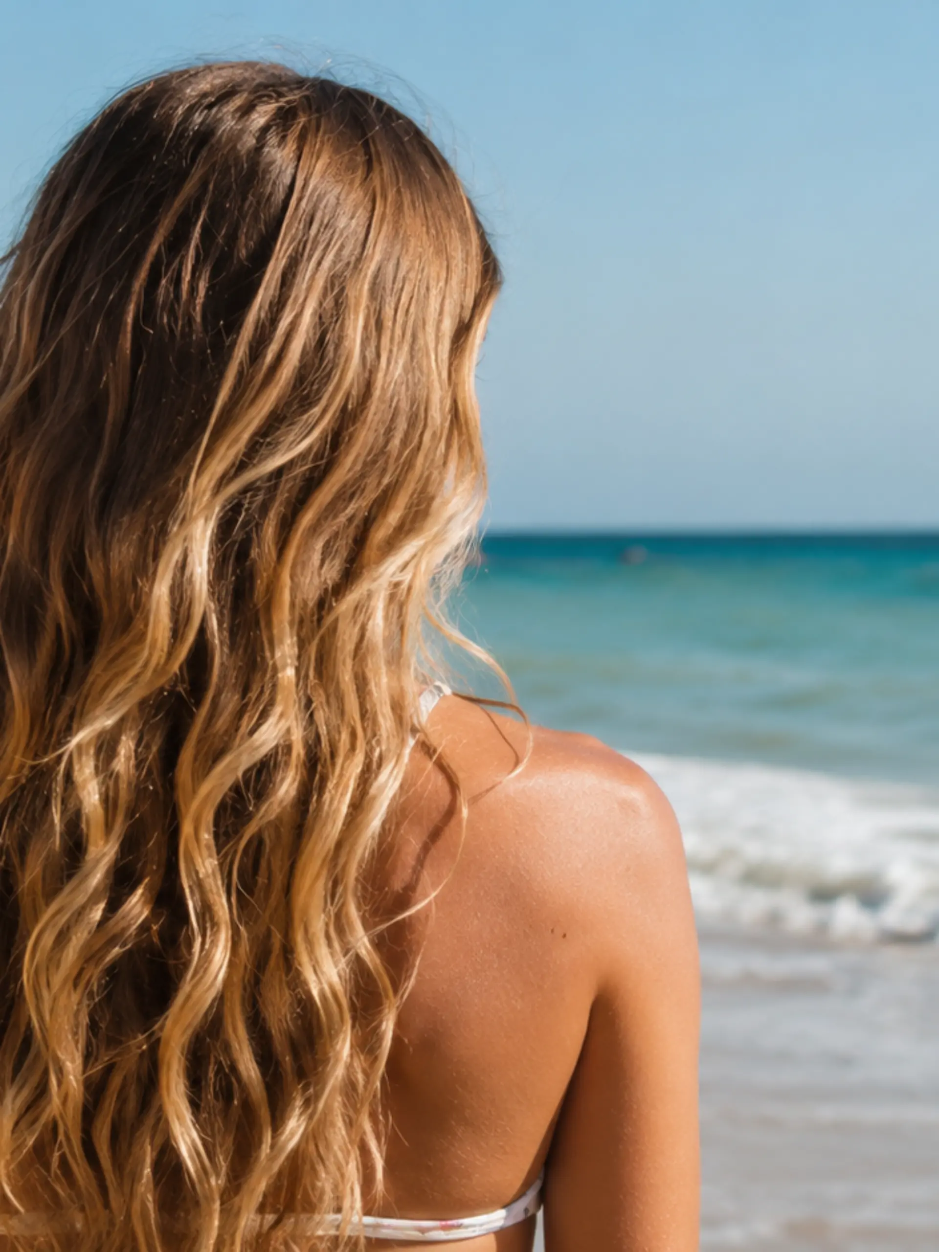 Frau mit langen, natürlichen Beach Waves steht von hinten am Strand und blickt aufs Meer – Symbolbild für den Frisurentrend Beach Waves 2026.