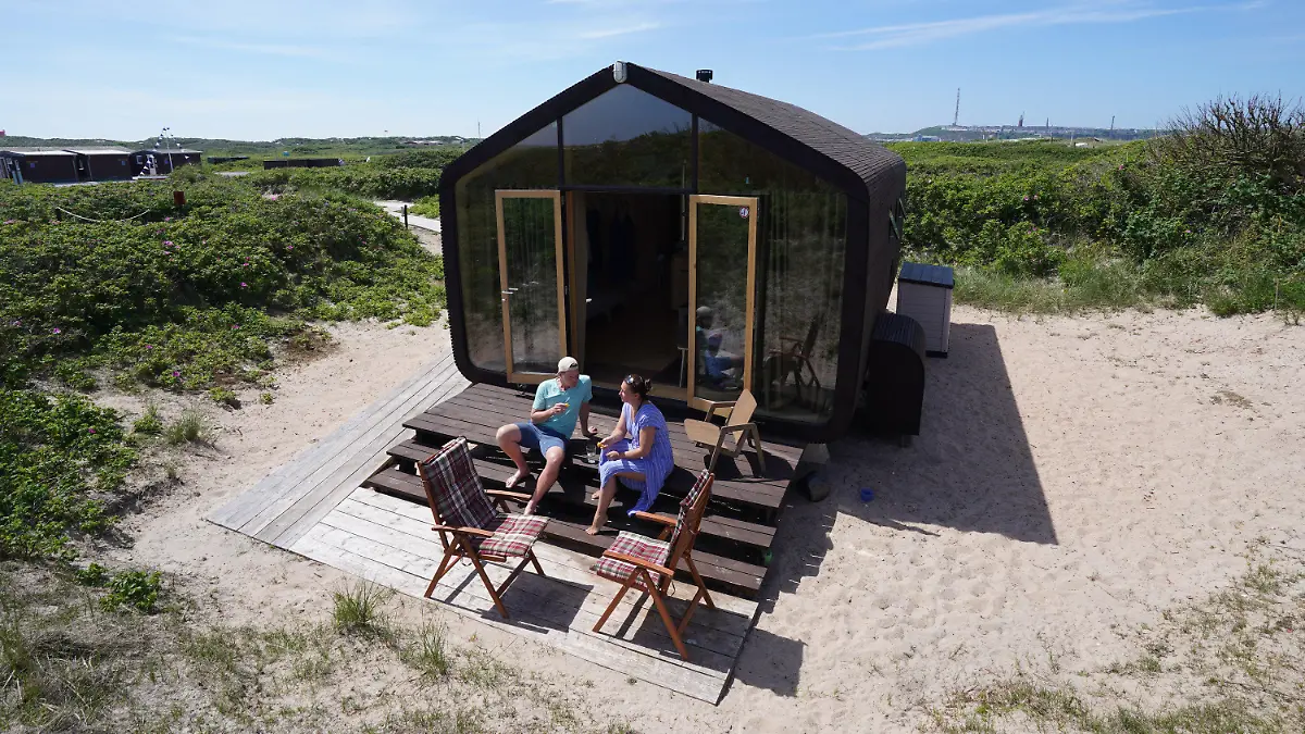 16.06.2021, Schleswig-Holstein, Helgoland: Monique Eichhorn und Sebastian Ebeling, Urlauber aus Berlin, sitzen auf den Stufen ihres gemieteten sogenannten Wikkelhouse auf der Helgoländer Düne. Die beiden sogenannten Wikkelhouses aus einer Manufaktur in Amsterdam sind nach Angaben des Helgoland Tourismus-Services für diese Saison ausgebucht. Auch im kommenden Jahr sind die Tiny Houses, die nur online und wochenweise gebucht werden können, bereits belegt. Die kleinen Häuser, deren Wände im Kern aus 24 miteinander verwickelter Schichten Pappe bestehen, sind komplett ausgestattet - es gibt aber kein Wasser und keine Toilette. (zu dpa «Gefragte Pappe: Helgoland möchte mehr Wikkelhouses») Foto: Marcus Brandt/dpa +++ dpa-Bildfunk +++