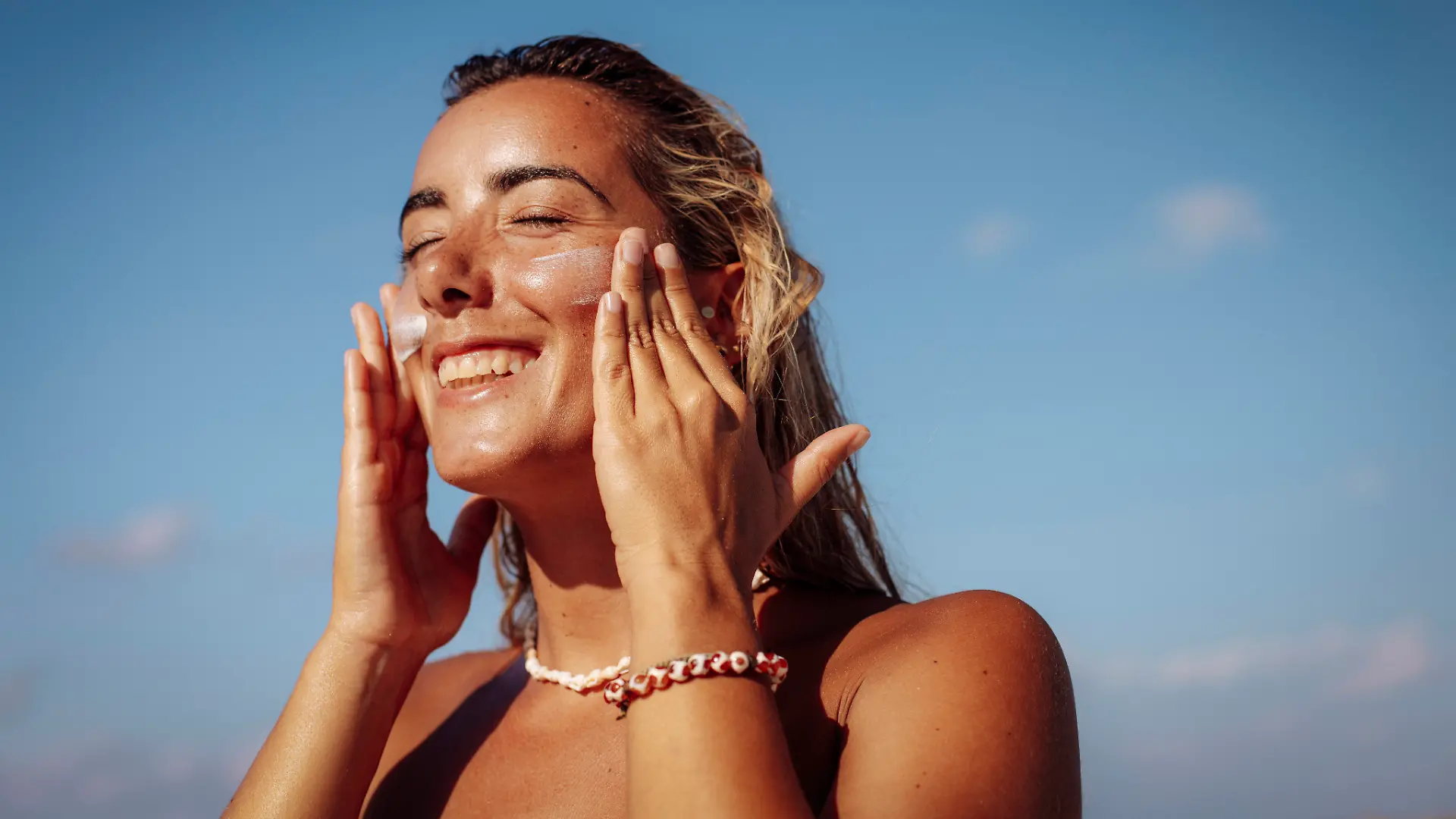 Frau am Strand lacht und trägt sich Sonnencreme im Gesicht auf