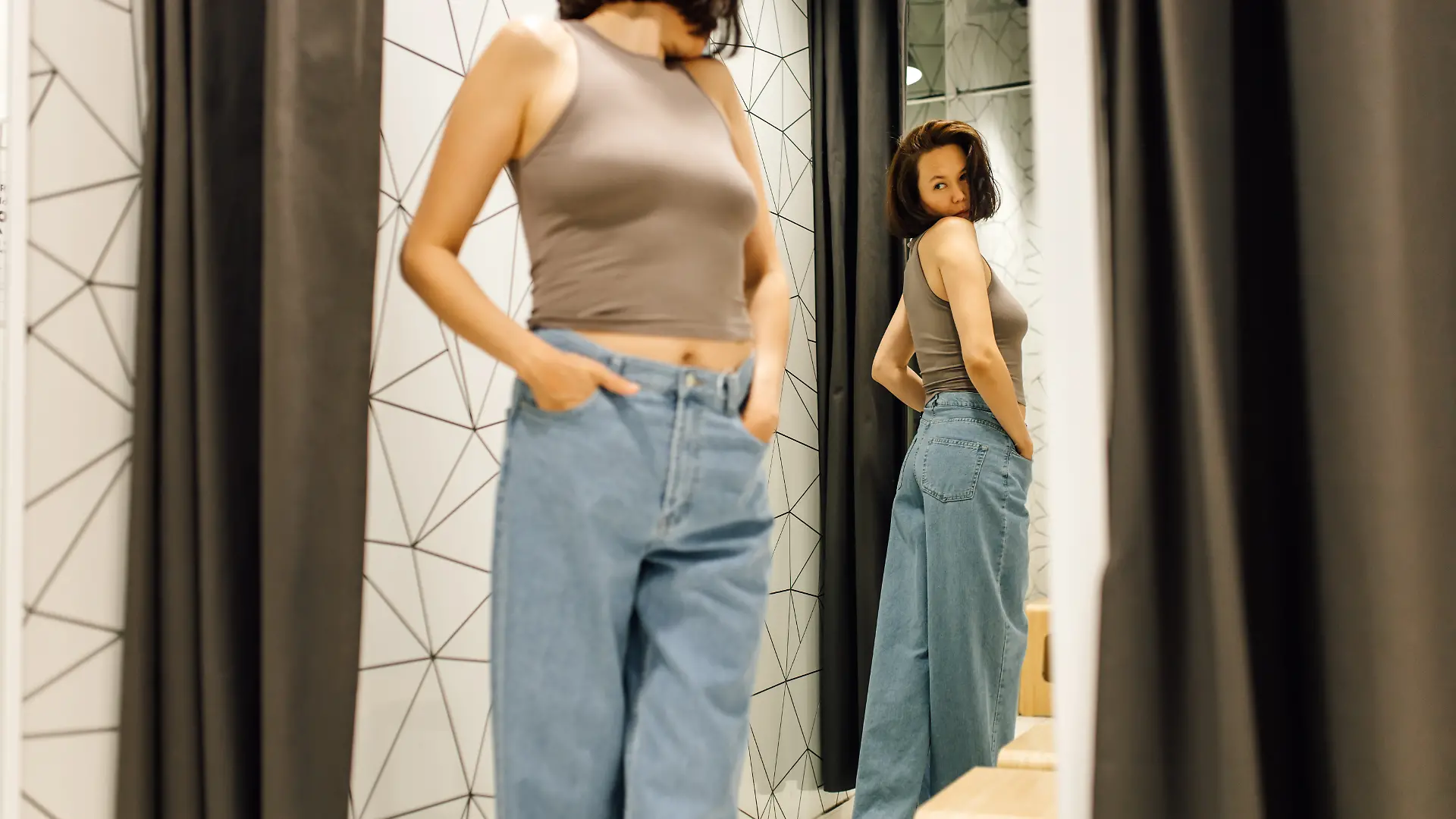 Eine Frau mit braunen Haaren betrachtet sich im Spiegel der Umkleidekabine, während sie eine neue Jeans und ein beiges Oberteil anprobiert.