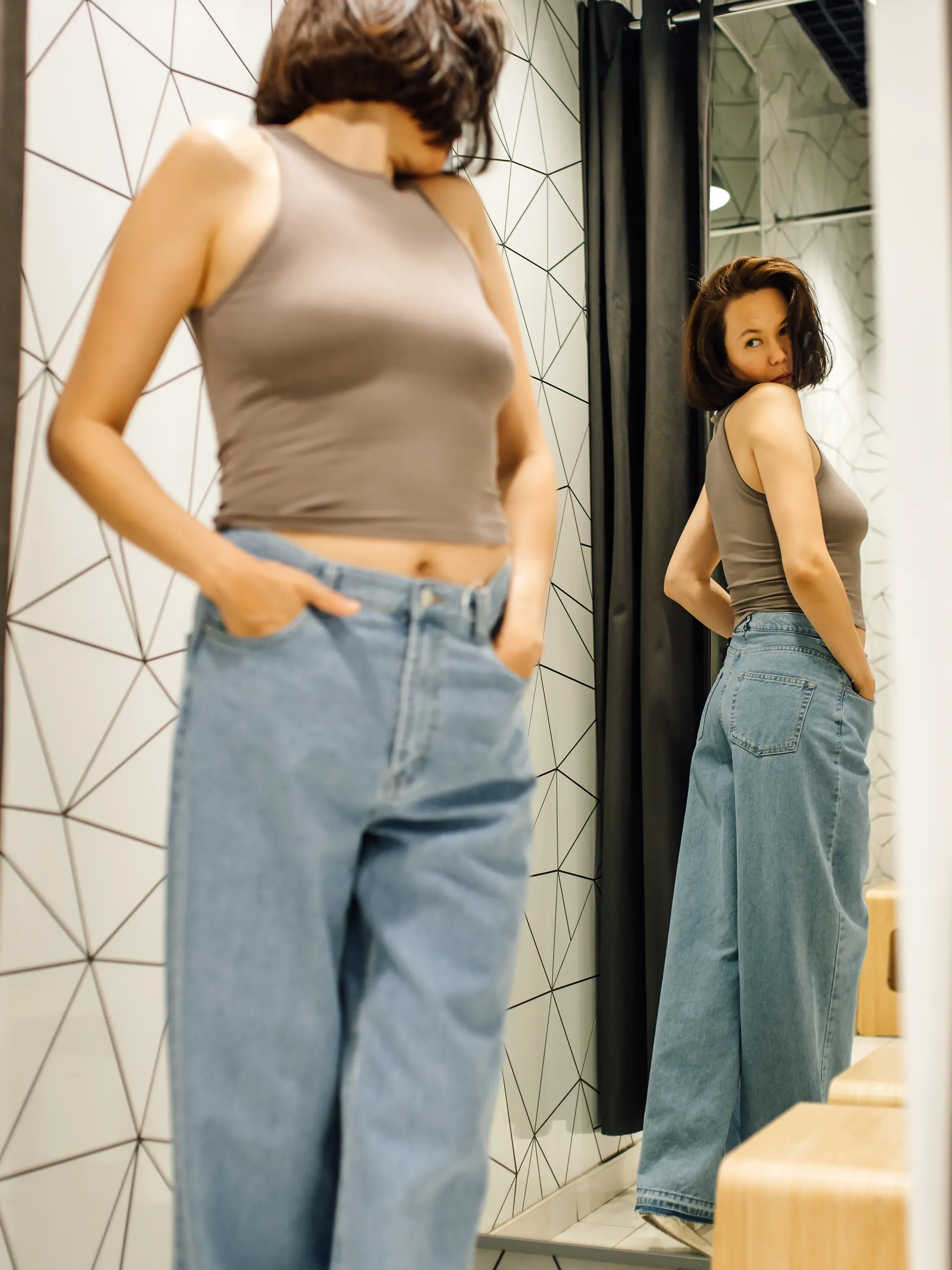 Eine Frau mit braunen Haaren betrachtet sich im Spiegel der Umkleidekabine, während sie eine neue Jeans und ein beiges Oberteil anprobiert.