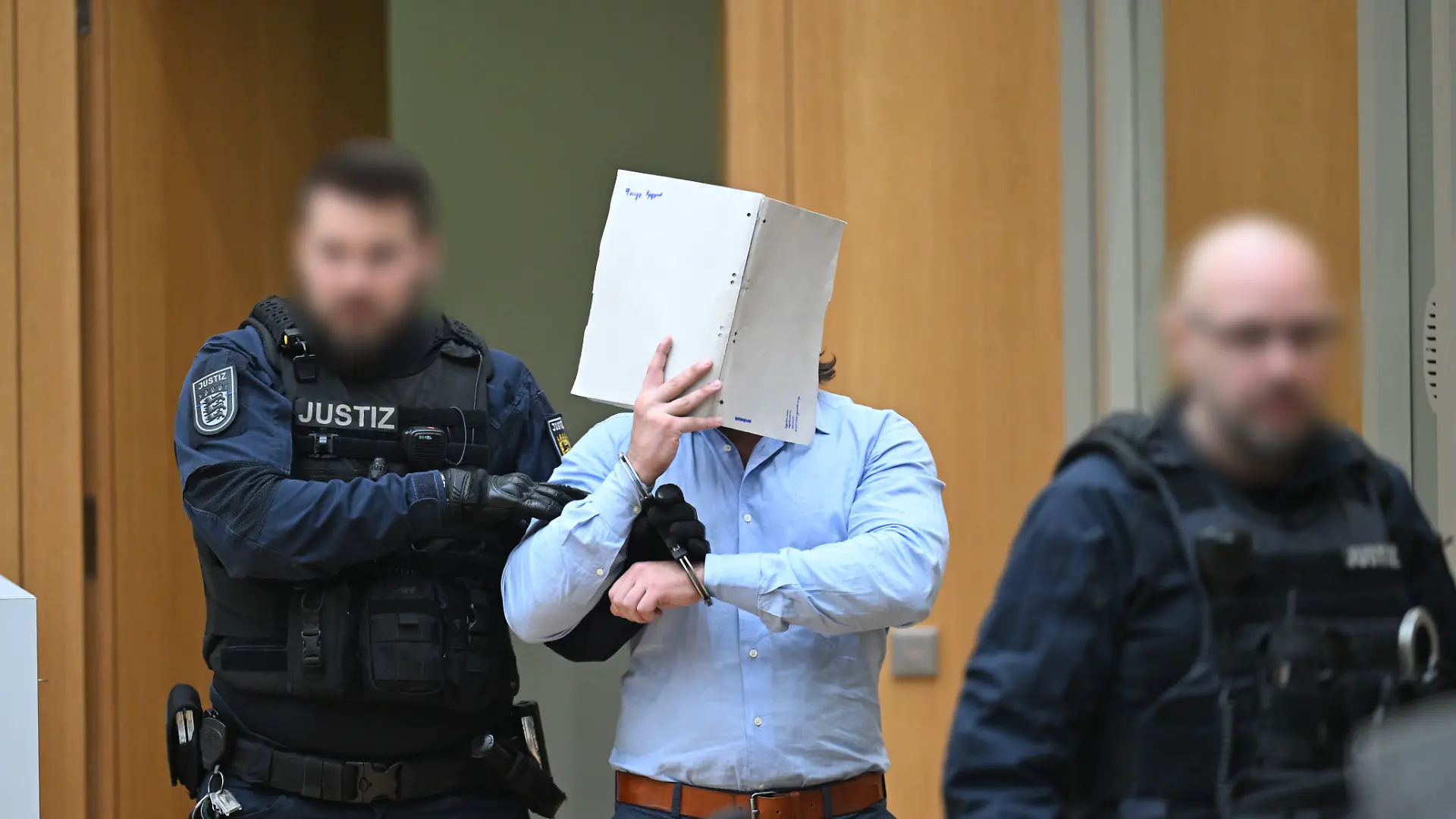 Einer der Angeklagten (M) in einem Staatsschutzverfahren gegen drei Ukrainer wegen Agententätigkeit im Auftrag eines russischen Nachrichtendienstes wird von Justizbeamten in den Saal des Oberlandesgerichts Stuttgart in Stammheim geführt. Die ukrainischen Angeklagten sollen im März 2025 über einen ukrainischen Postdienstleister zwei Pakete mit GPS-Trackern in Richtung Ukraine versandt haben, um Versandrouten und Transportabläufe bei dem Paketdienstleister auszuforschen.