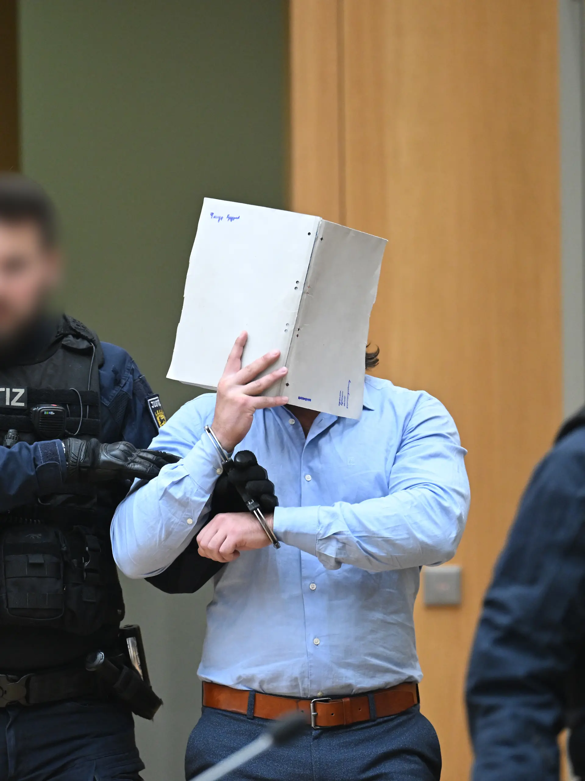 Einer der Angeklagten (M) in einem Staatsschutzverfahren gegen drei Ukrainer wegen Agententätigkeit im Auftrag eines russischen Nachrichtendienstes wird von Justizbeamten in den Saal des Oberlandesgerichts Stuttgart in Stammheim geführt. Die ukrainischen Angeklagten sollen im März 2025 über einen ukrainischen Postdienstleister zwei Pakete mit GPS-Trackern in Richtung Ukraine versandt haben, um Versandrouten und Transportabläufe bei dem Paketdienstleister auszuforschen.