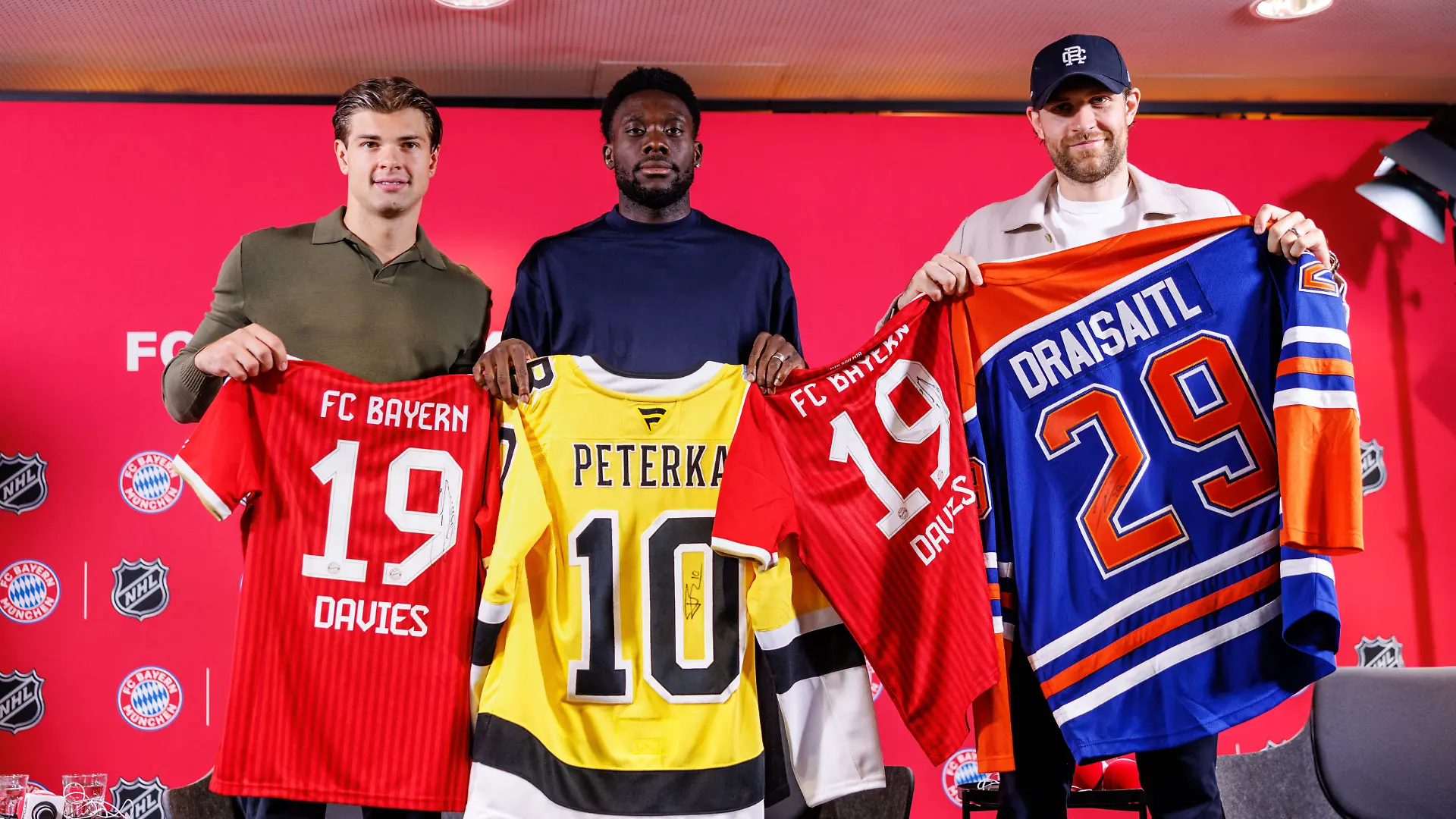 Eishockey, Fußball: Gemeinsame Pk des FC Bayern München und der National Hockey League (NHL). John-Jason Peterka (l-r), Spieler der Boston Bruins, Alphonso Davies, Spieler vom FC Bayern München, und Leon Draisaitl, Spieler der Edmonton Oilers, posieren auf der Pressekonferenz mit Trikots.