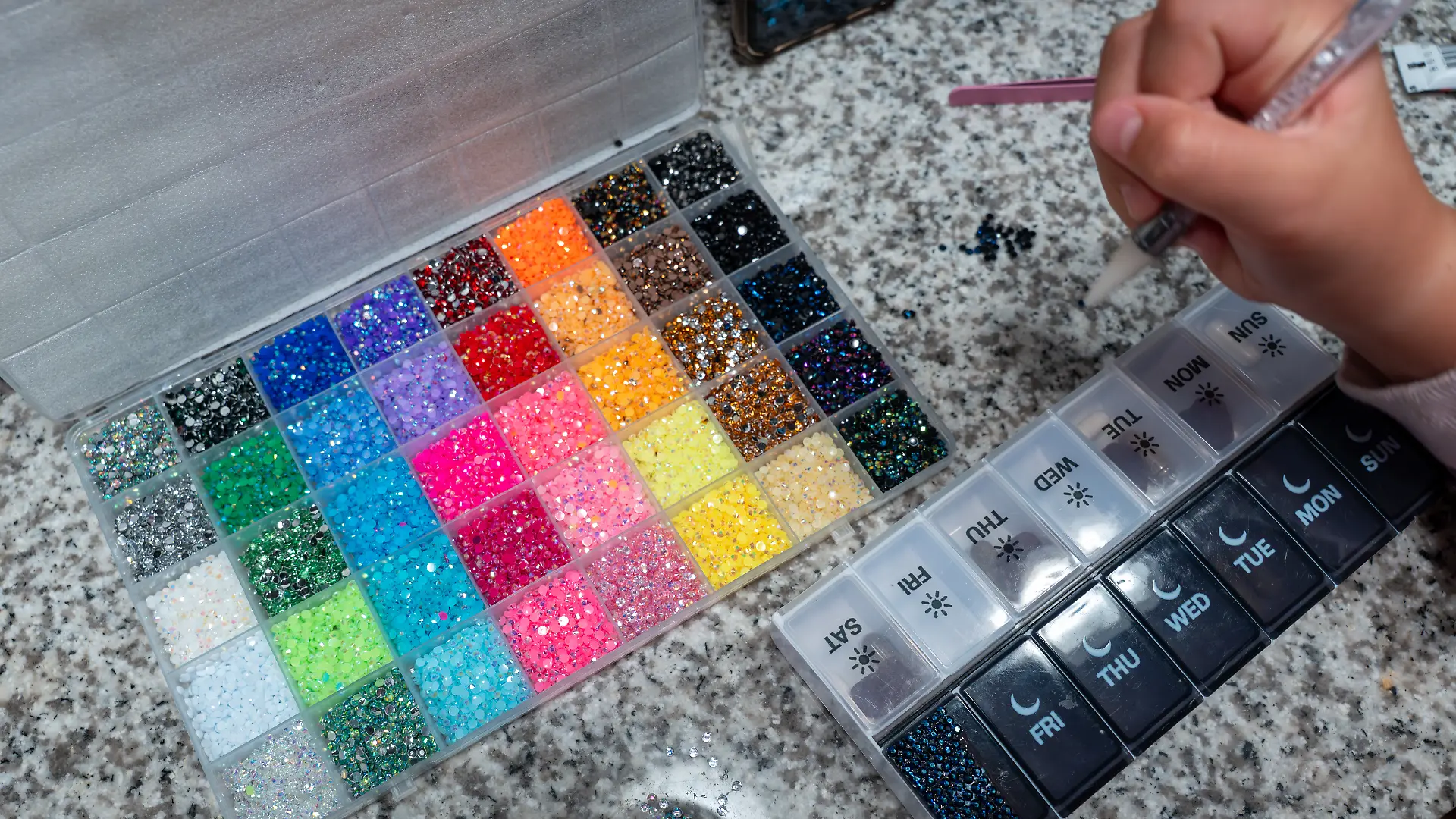 Kinderhand hält Glitzerstift in der Hand, umgeben von einer Box, gefüllt mit vielen bunten Glitzersteinchen