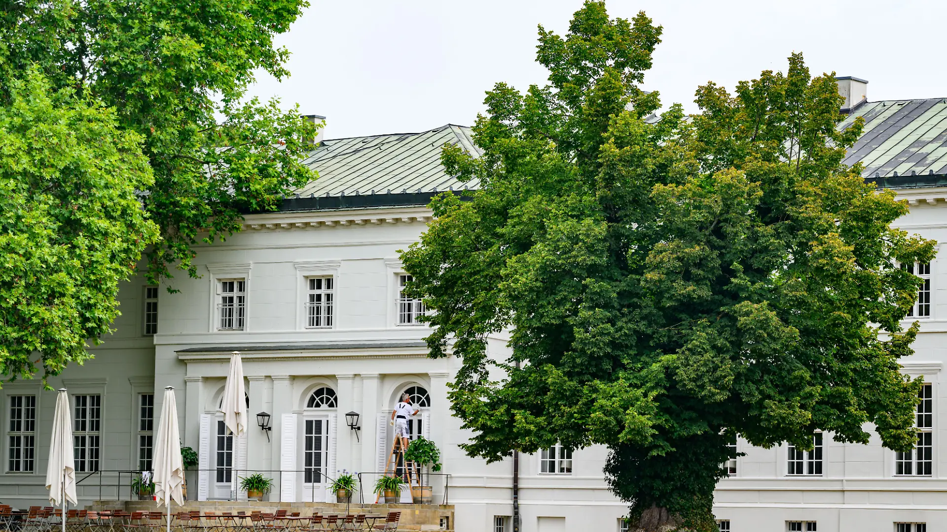 Ein Handwerker steht auf einer Leiter und verrichtet Malerarbeiten an einem Fenster der weißen Schlossfassade, die sich hinter einer Außengastronomie mit geschlossenen Sonnenschirmen und einem mächtigen, alten Laubbaum erstreckt. Das historische Bauwerk wurde Anfang des 19. Jahrhunderts nach Entwürfen des berühmten Baumeisters Karl Friedrich Schinkel im Stil des Klassizismus umfassend umgestaltet. Heute dient die geschichtsträchtige Anlage als renommiertes Kulturzentrum für Ausstellungen, Konferenzen und hochkarätige Veranstaltungen im ländlichen Raum. Die erhaltene Architektur mit ihren klaren Symmetrien und historischen Details zieht ganzjährig kulturinteressierte Besucher und Touristen an.