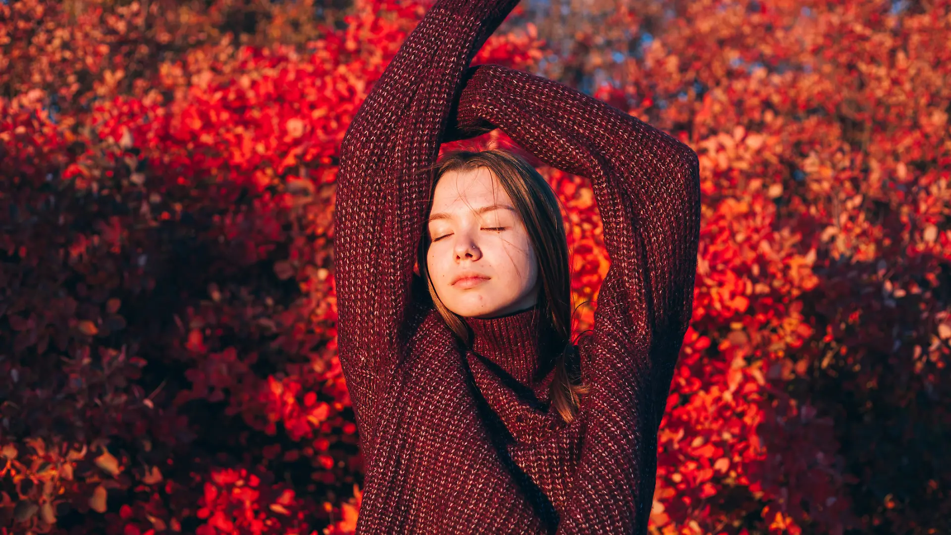Eine Frau streckt sich vor einem herbstlichen Baum und trägt einen bordeauxroten Strickpullover.