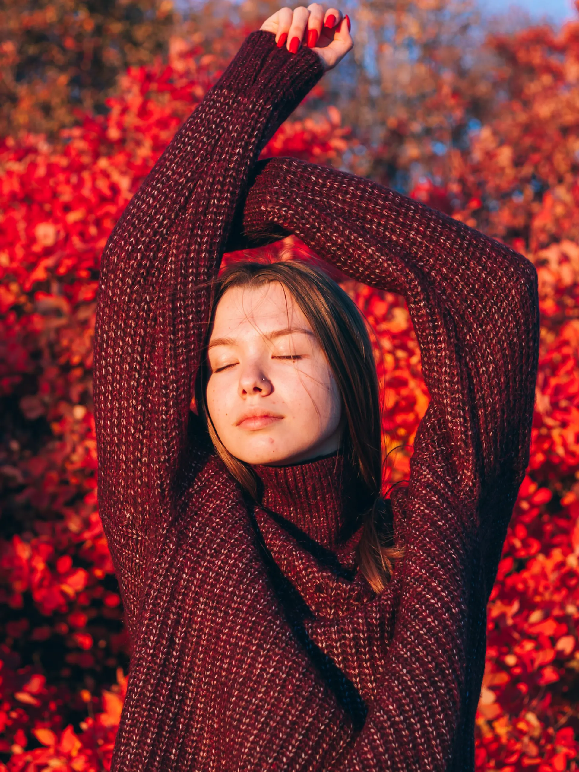 Eine Frau streckt sich vor einem herbstlichen Baum und trägt einen bordeauxroten Strickpullover.