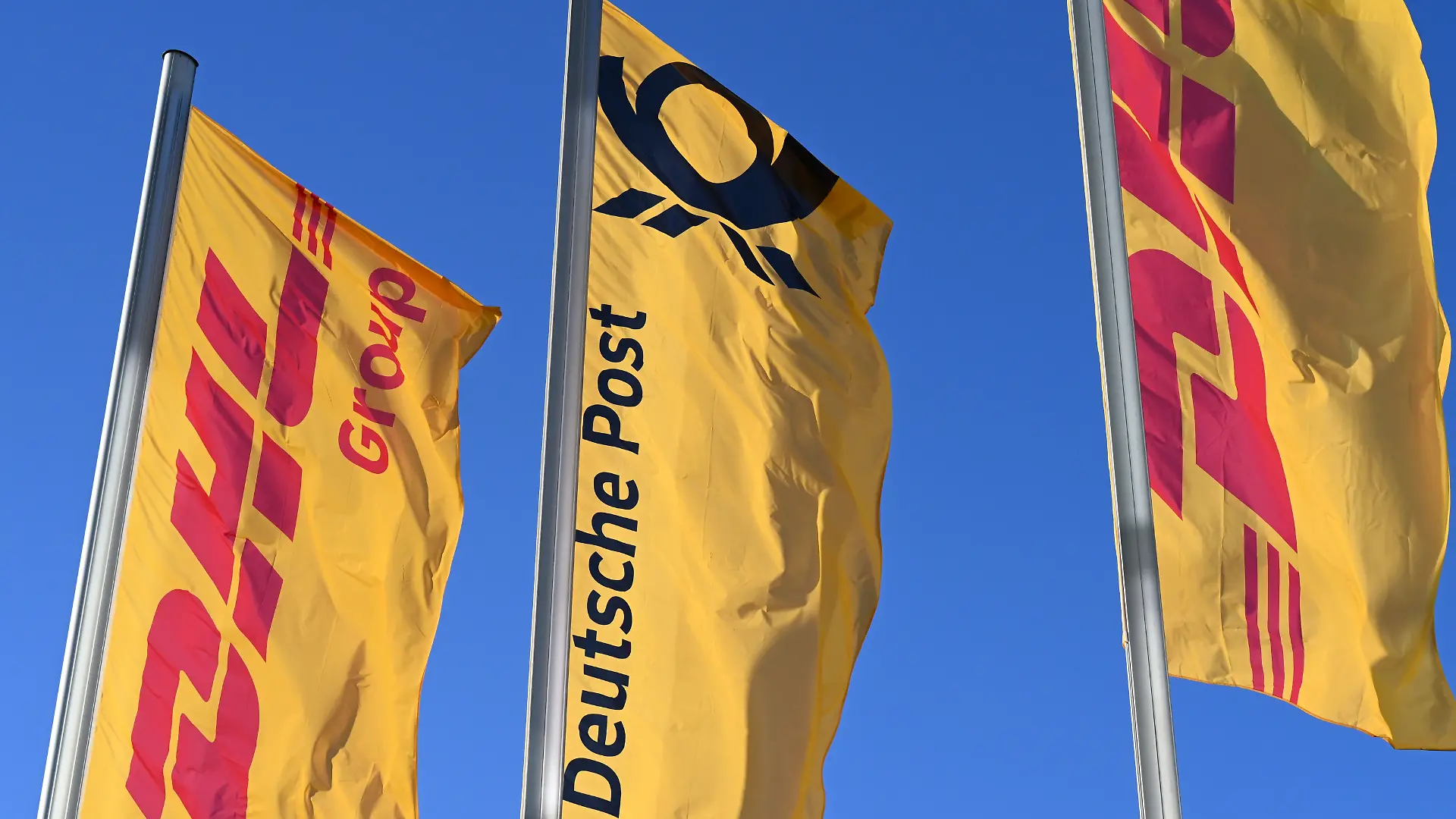 ARCHIV - 13.01.2025, Nordrhein-Westfalen, Bonn: Fahnen des Logistikkonzerns DHL und seiner Brieftochter Deutsche Post (M) wehen vor der Konzernzentrale an Masten. Der Konzern hat sich von Deutsche Post AG zu DHL AG umbenannt. (zu dpa: «Bonner Logistikkonzern heißt jetzt DHL statt Deutsche Post») Foto: Wolf von Dewitz/dpa +++ dpa-Bildfunk +++
