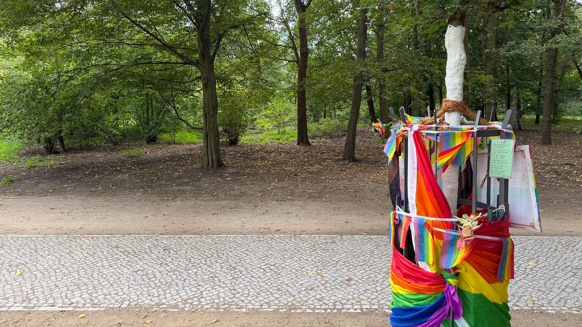 Die zerstörte Regenbogenbank wurde inzwischen abgebaut. Gegenüber steht der neu gepflanzte Baum, der ebenfalls an den CSD-Anschlag erinnern soll.