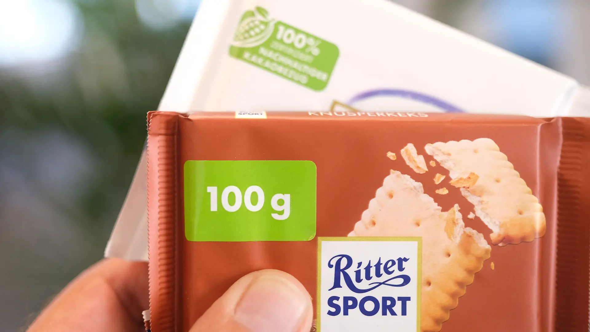 Eine Hand hält zwei Tafeln Schokolade von Ritter Sport. Auf der vorderen Tafel ist auf der Verpackung ein Hinweis mit einem Sticker überklebt. (zu dpa: «EU-Richtlinie zwingt Ritter Sport zu neuen Verpackungen»)