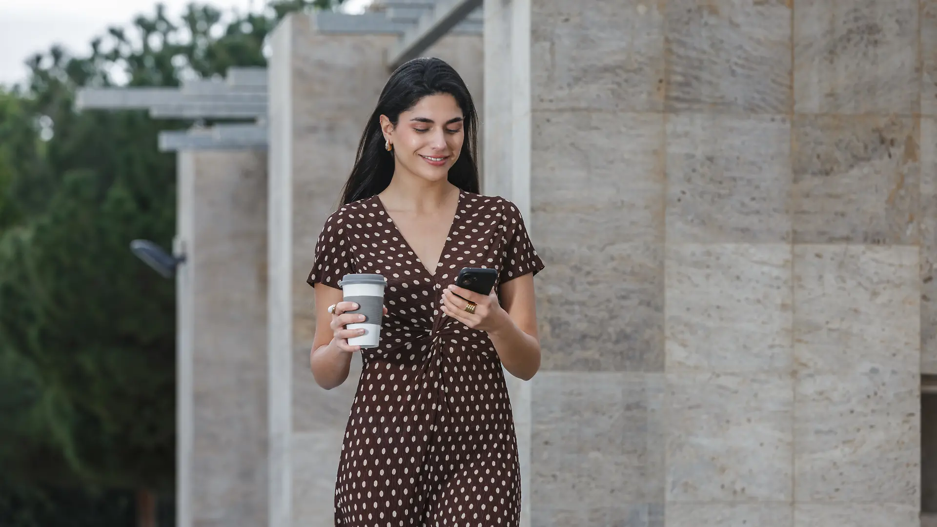 Frau mit dunklen Haaren läuft in einem braunen Kleid mit weißen Punkten auf dem Gehweg. In der einen Hand hat sie ein Smartphone, in der anderen einen To-go-Becher.