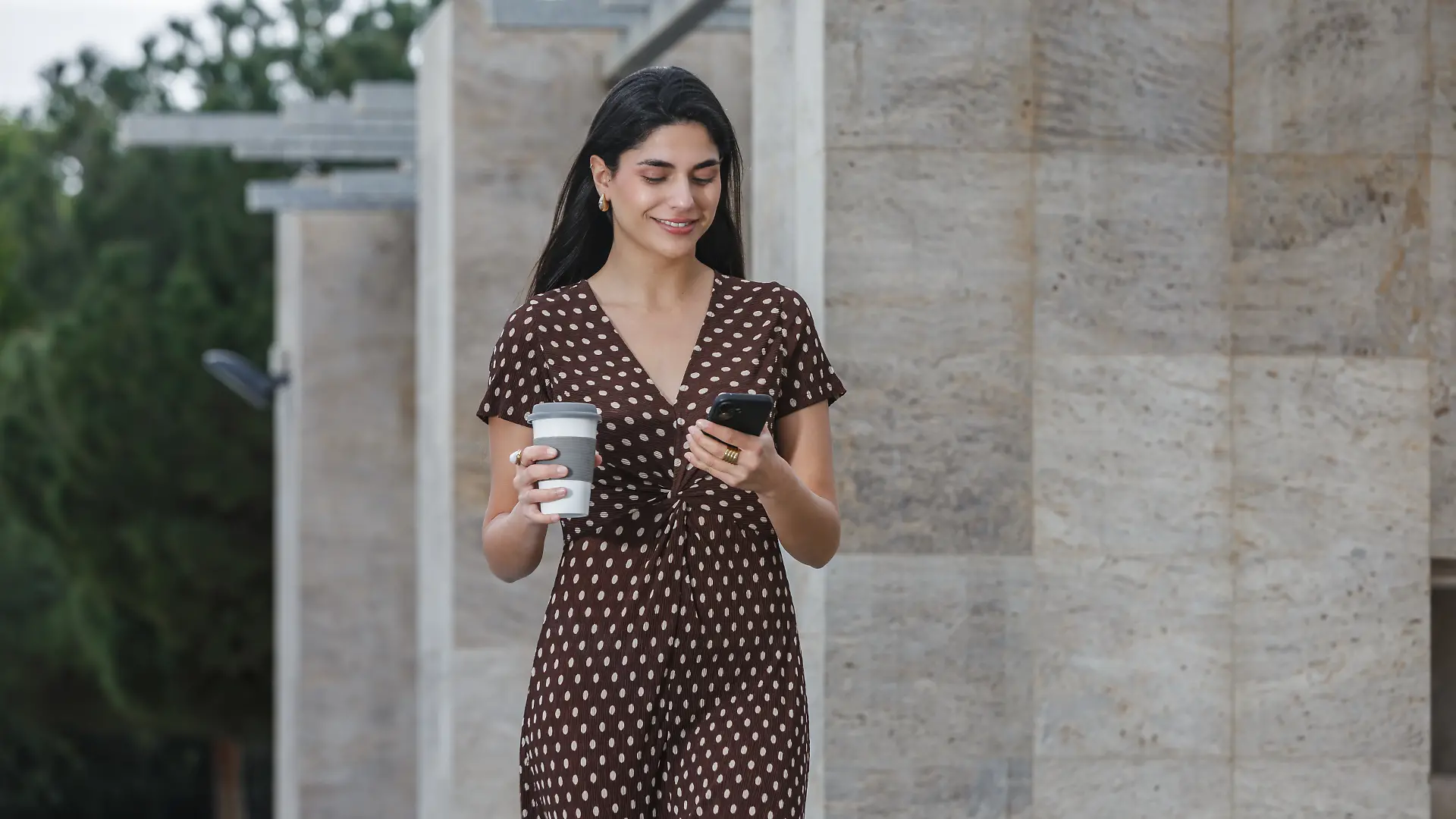Frau mit dunklen Haaren läuft in einem braunen Kleid mit weißen Punkten auf dem Gehweg. In der einen Hand hat sie ein Smartphone, in der anderen einen To-go-Becher.