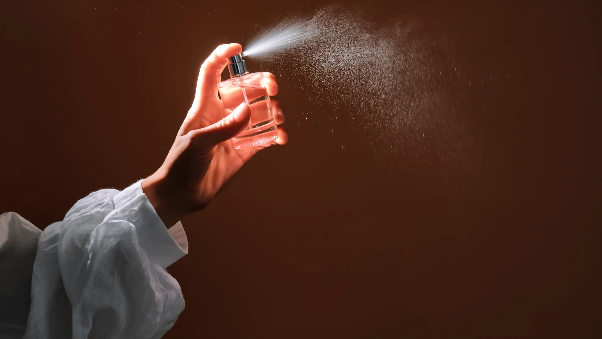 Die Hand einer Person, die Parfüm aus einer Glasflasche versprüht und dabei einen feinen Nebel erzeugt