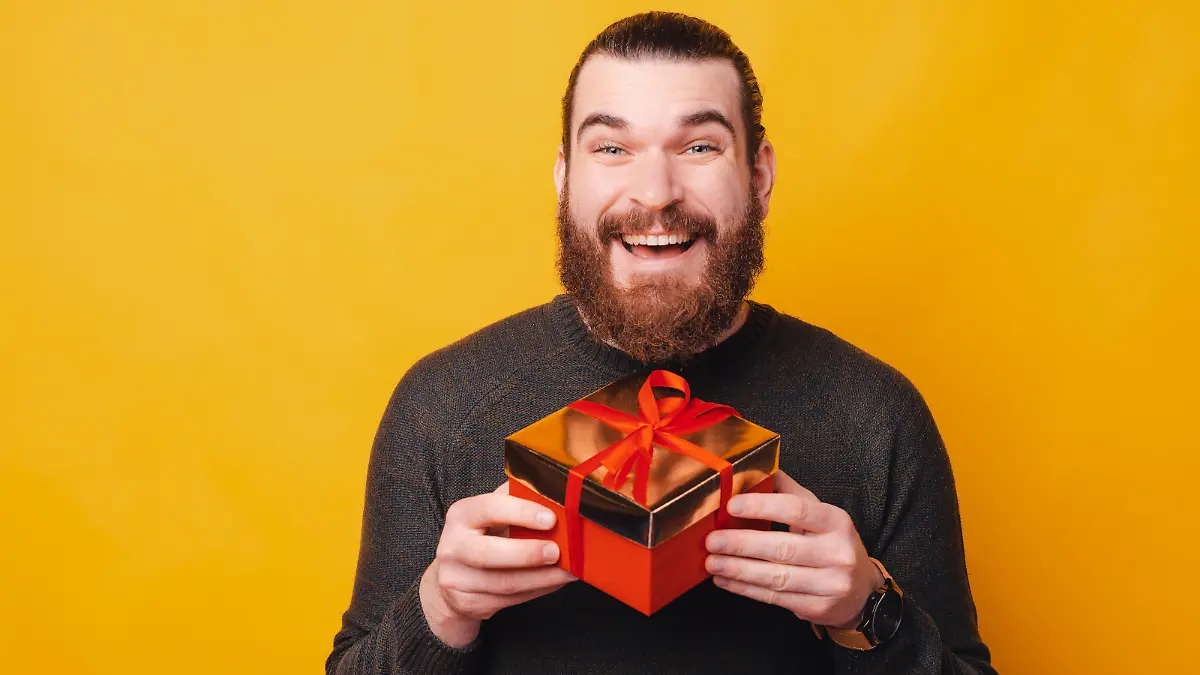 Charming young bearded man holding gift box over yellow background.