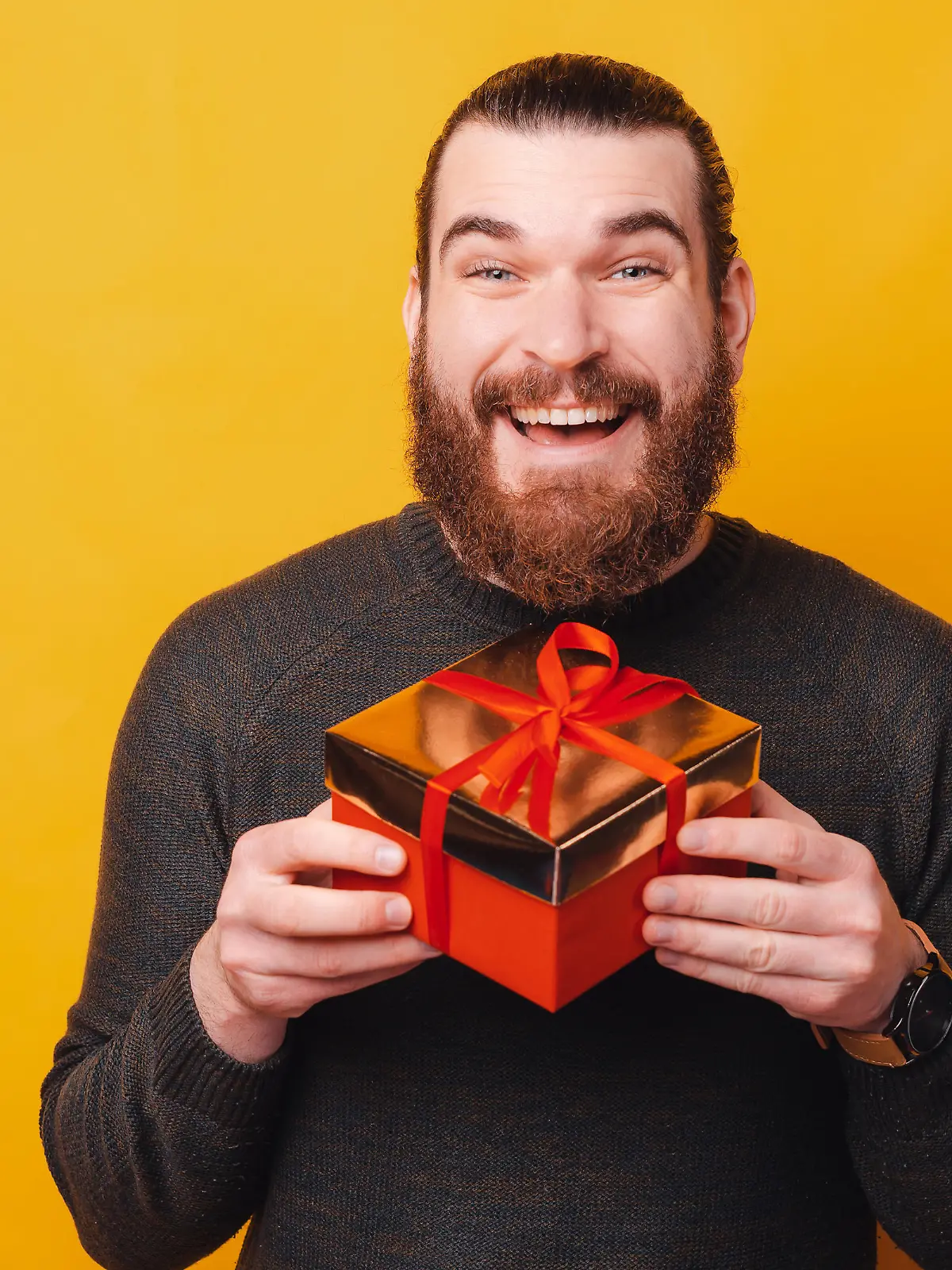 Charming young bearded man holding gift box over yellow background.