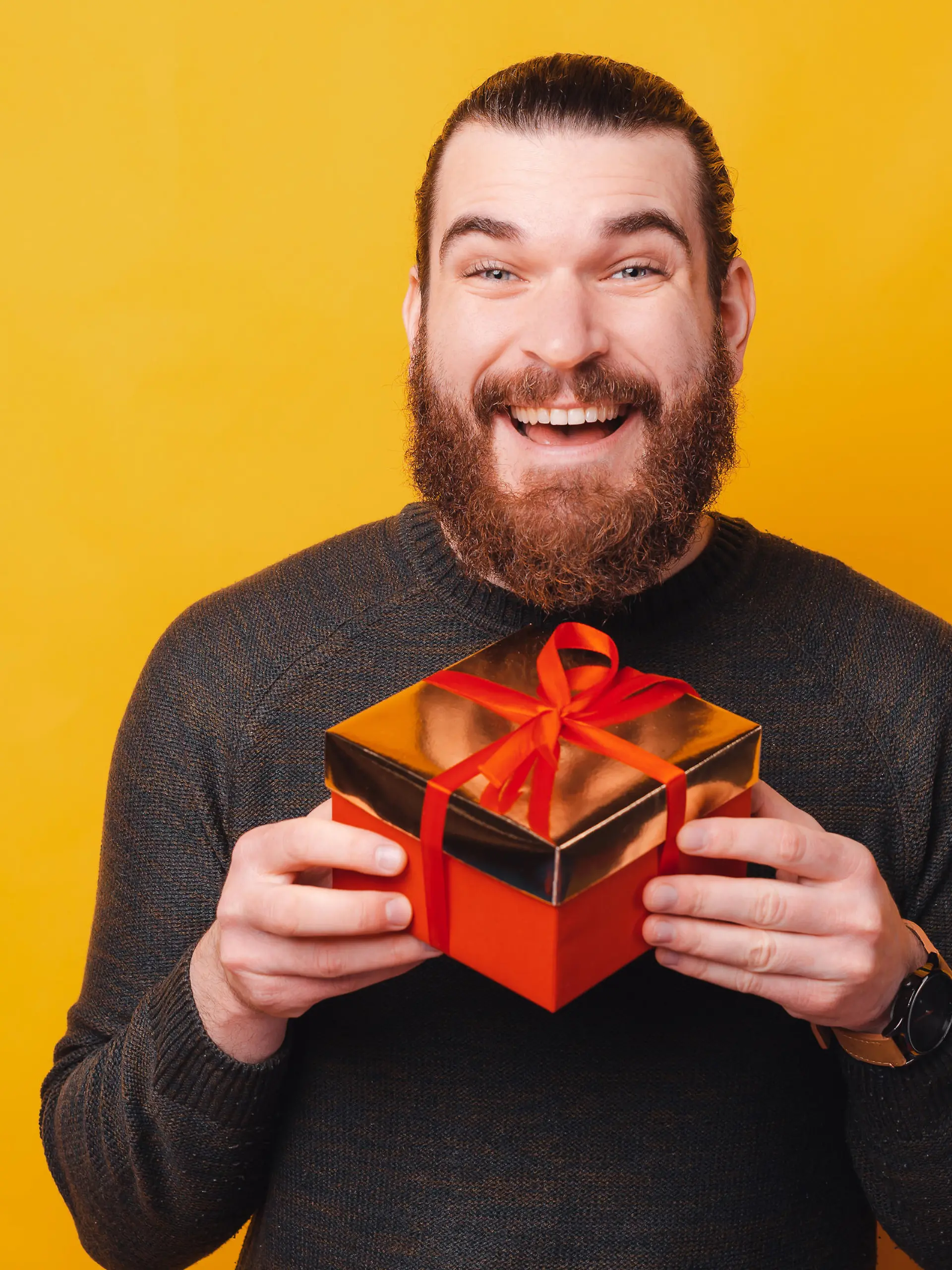 Charming young bearded man holding gift box over yellow background.