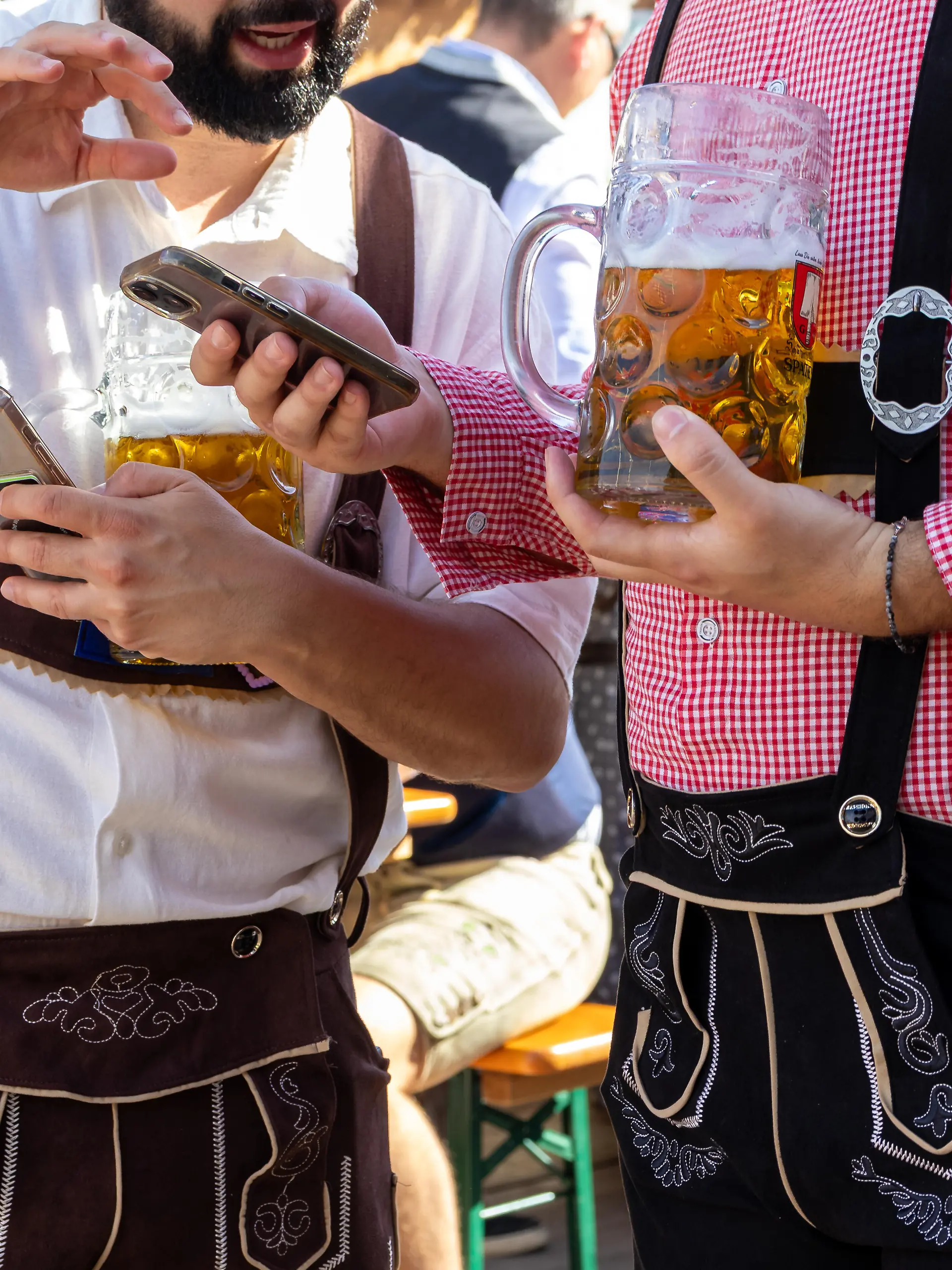 Zwei Besucher in Lederhosen halten Maßkrüge mit Bier und nutzen ihre Smartphones beim 191. Oktoberfest 2026. Tracht, Geselligkeit und digitale Kommunikation treffen aufeinander
