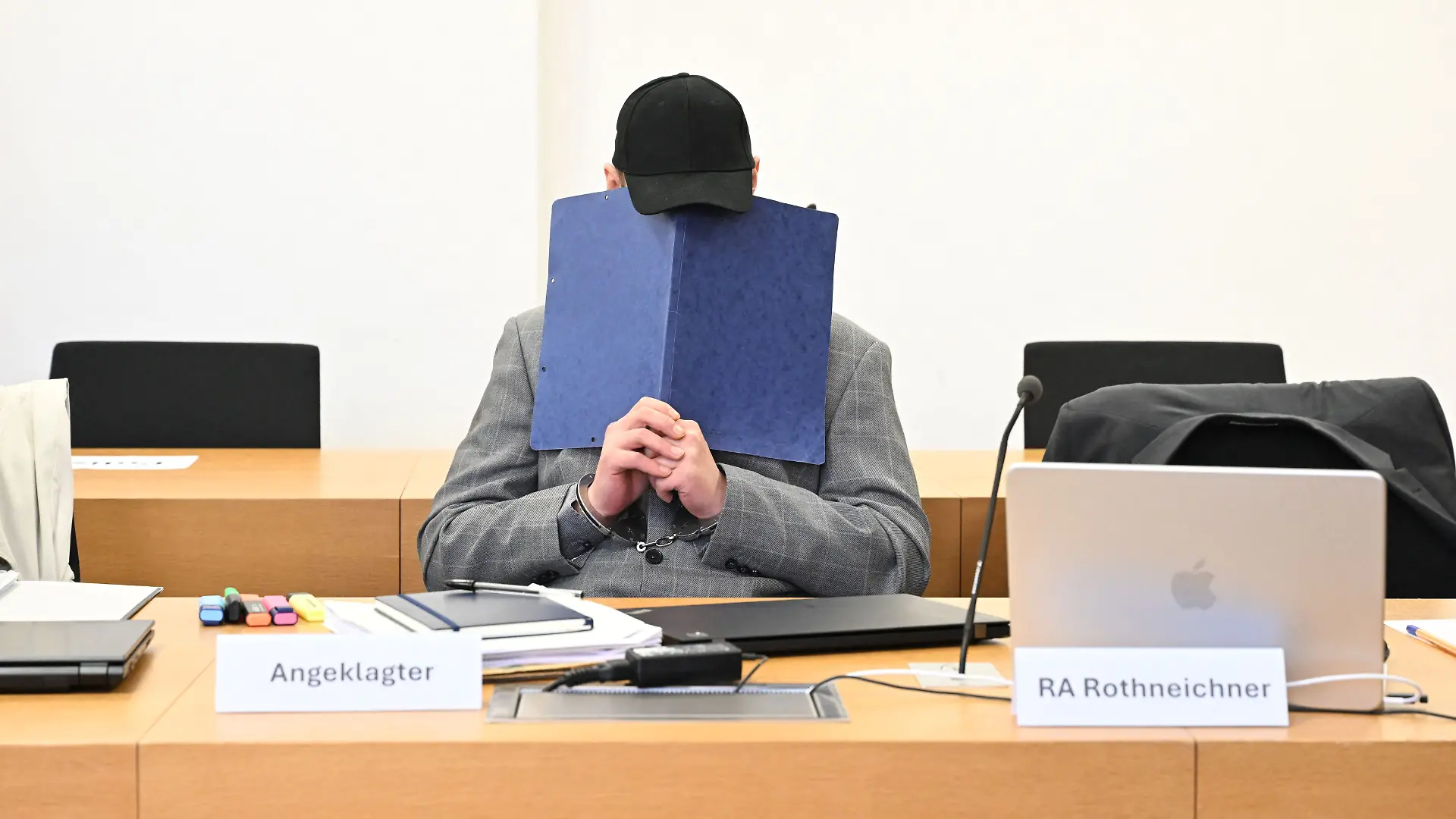 Zu Beginn eines Prozesses um einen Dreifachmord in Zwiesel sitzt der Angeklagte im Gerichtssaal des Landgerichtes Deggendorf und verdeckt sein Gesicht mit einem Aktendeckel. 