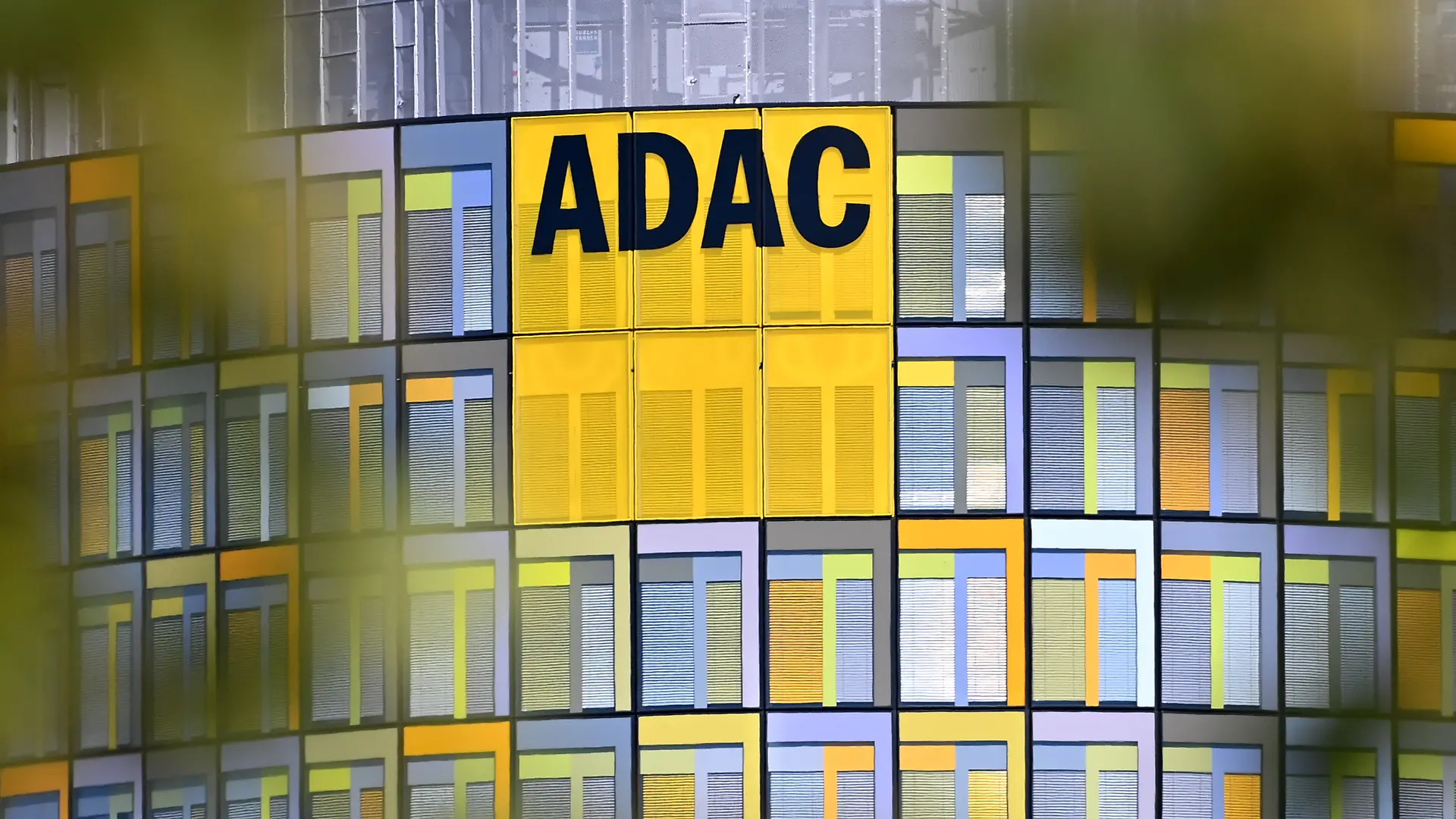 ADAC Hauptzentrale. Die moderne und farbenfrohe Glasfassade der ADAC-Zentrale in Muenchen mit dem markanten gelben Logo, eingerahmt von unscharfen Blaettern im Vordergrund. *** ADAC Headquarters: The modern and colorful glass facade of the ADAC headquarters in Munich, featuring the striking yellow logo, framed by out-of-focus leaves in the foreground