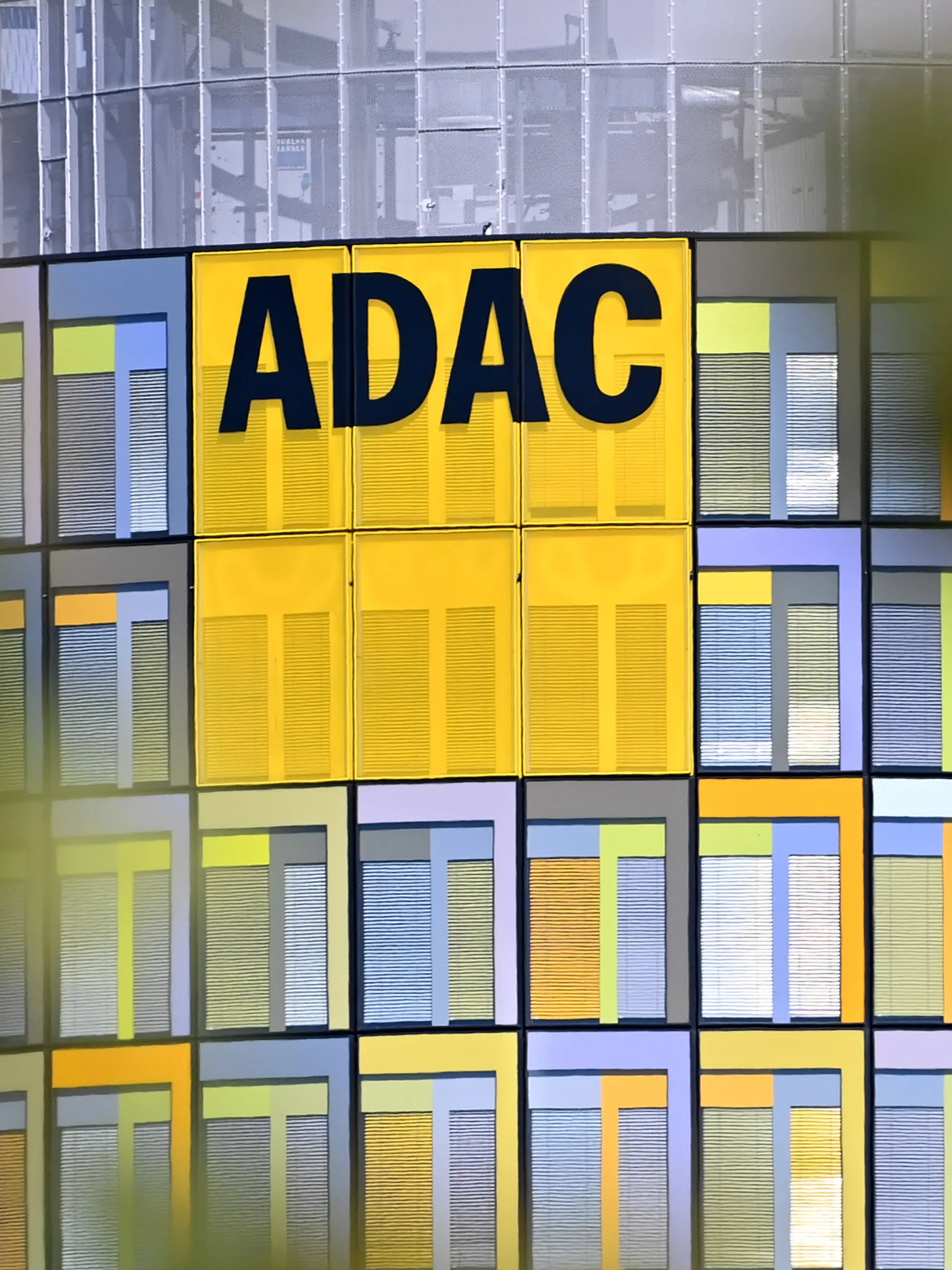 ADAC Hauptzentrale. Die moderne und farbenfrohe Glasfassade der ADAC-Zentrale in Muenchen mit dem markanten gelben Logo, eingerahmt von unscharfen Blaettern im Vordergrund. *** ADAC Headquarters: The modern and colorful glass facade of the ADAC headquarters in Munich, featuring the striking yellow logo, framed by out-of-focus leaves in the foreground