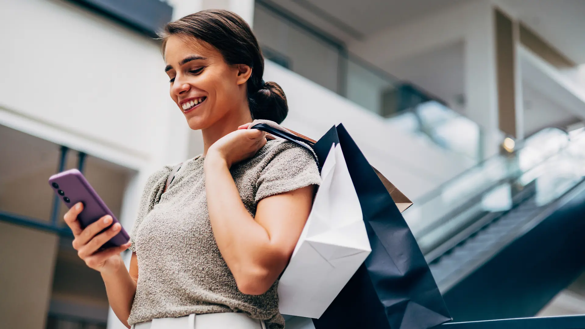 Eine Frau mit Shopping-Bags auf der Schulter schaut in einem Einkaufszentrum auf ihr Smartphone.