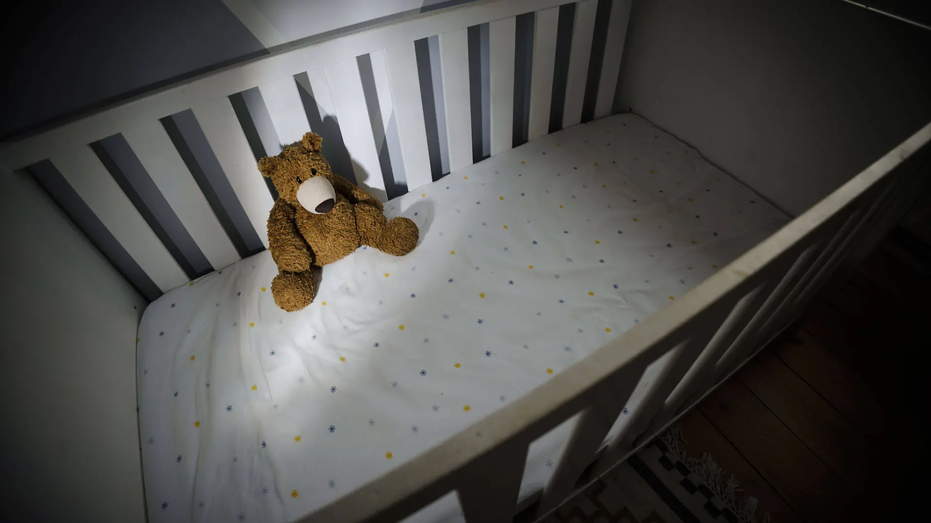 Ein Teddybaer sitzt in einem leeren Kinderbett. 