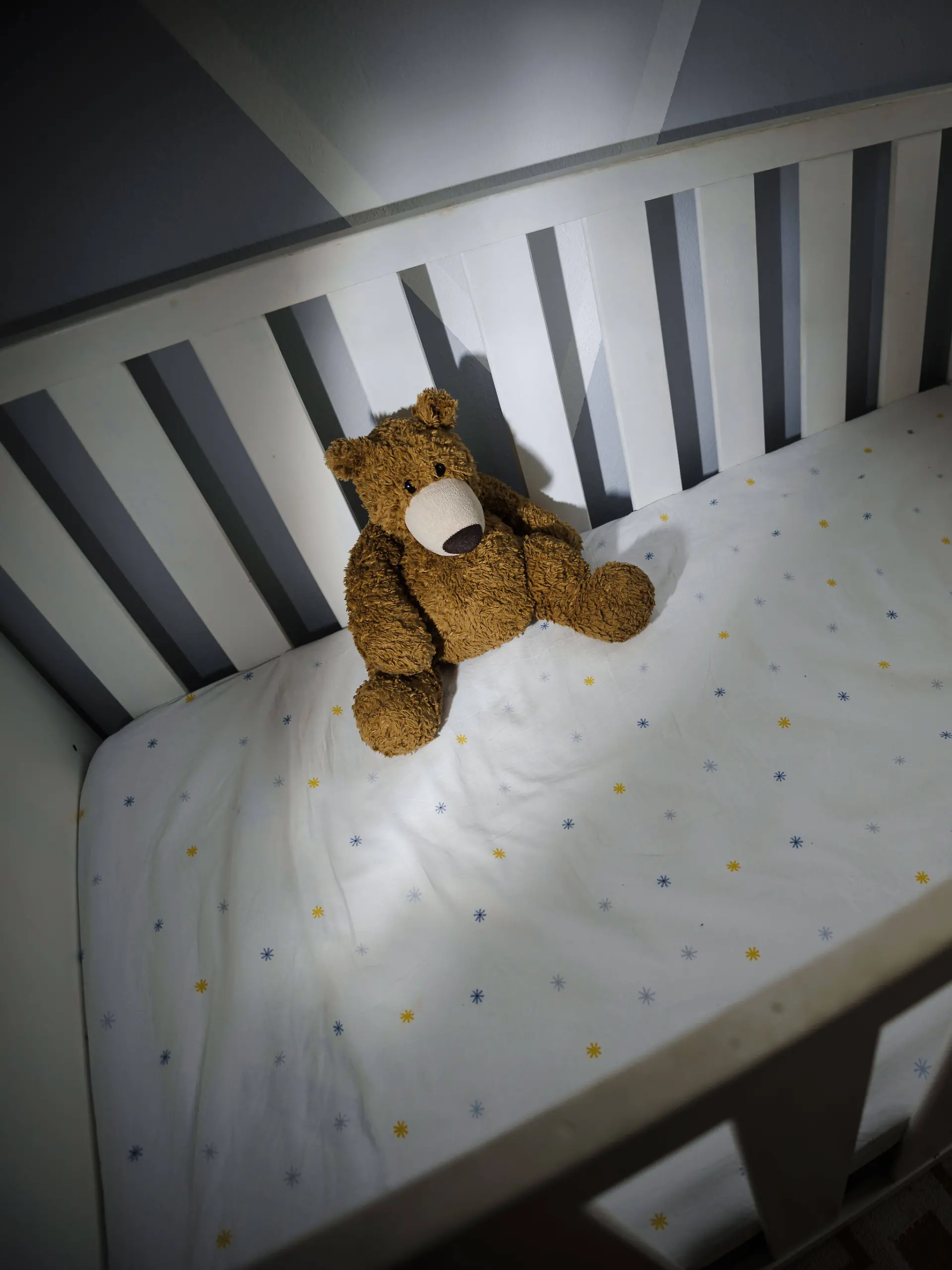 Ein Teddybaer sitzt in einem leeren Kinderbett. 