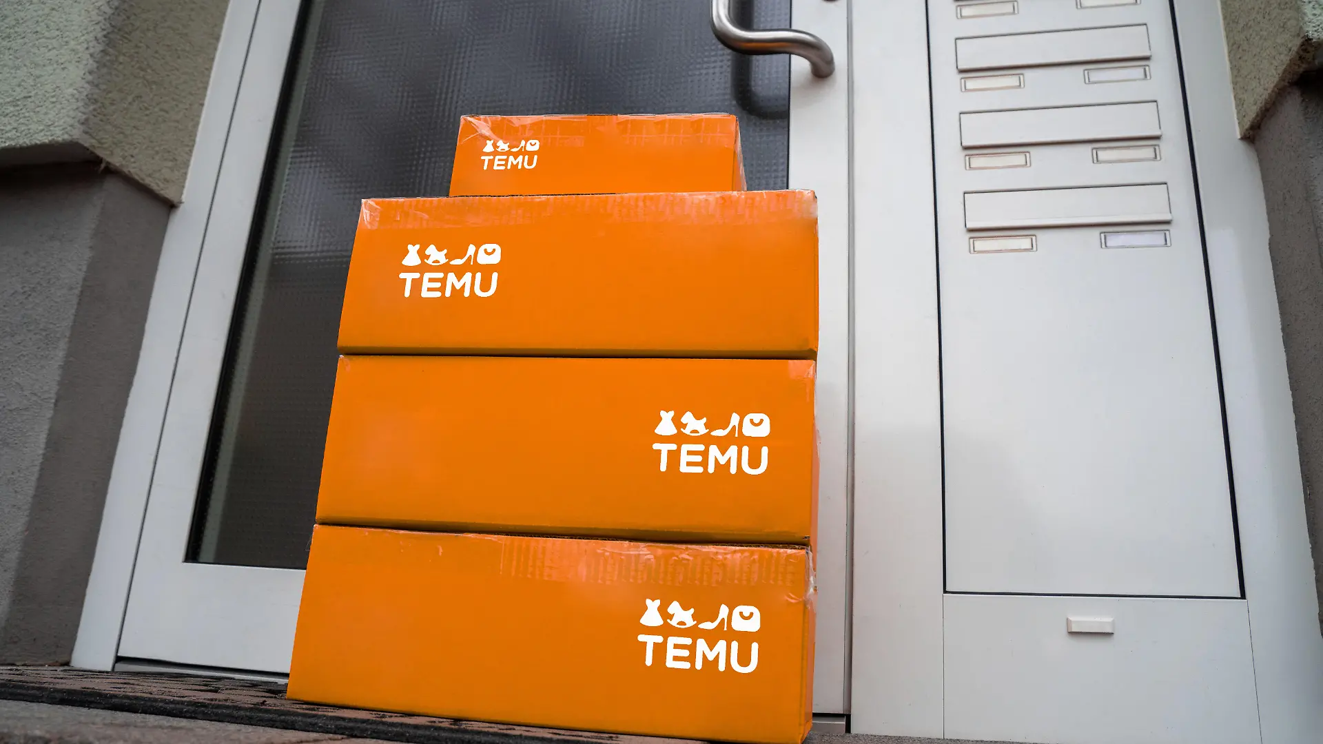 Symbolbild: Orangefarbene Temu-Pakete vor einer Haustür verdeutlichen den Boom von internationalen E-Commerce-Plattformen. Temu, bekannt für günstige Preise und große Produkt-Auswahl, aggressive Marketingstrategien in Europa und den USA. Themen wie nachhaltige Verpackung, Lieferzeiten und Zollprobleme sind dabei zentral für den Erfolg solcher Plattformen. FOTOMONTAGE
