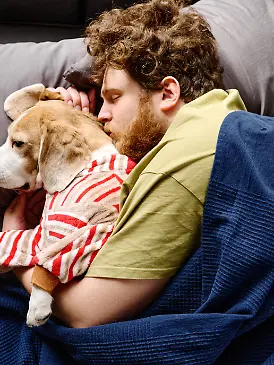 High angle view of bearded young man lying under blanket in bed and cuddling his dog || Modellfreigabe vorhanden