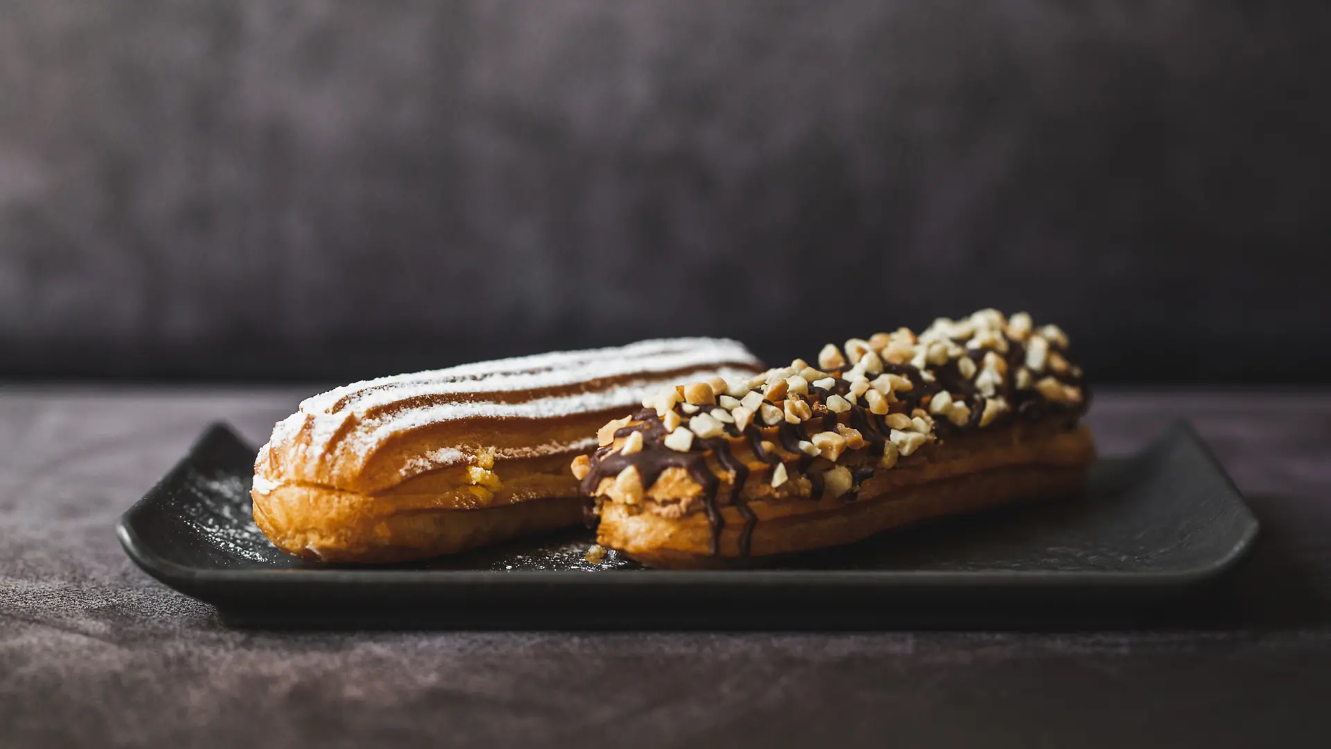 Zwei Eclairs auf einem Teller vor dunklem Hintergrund.