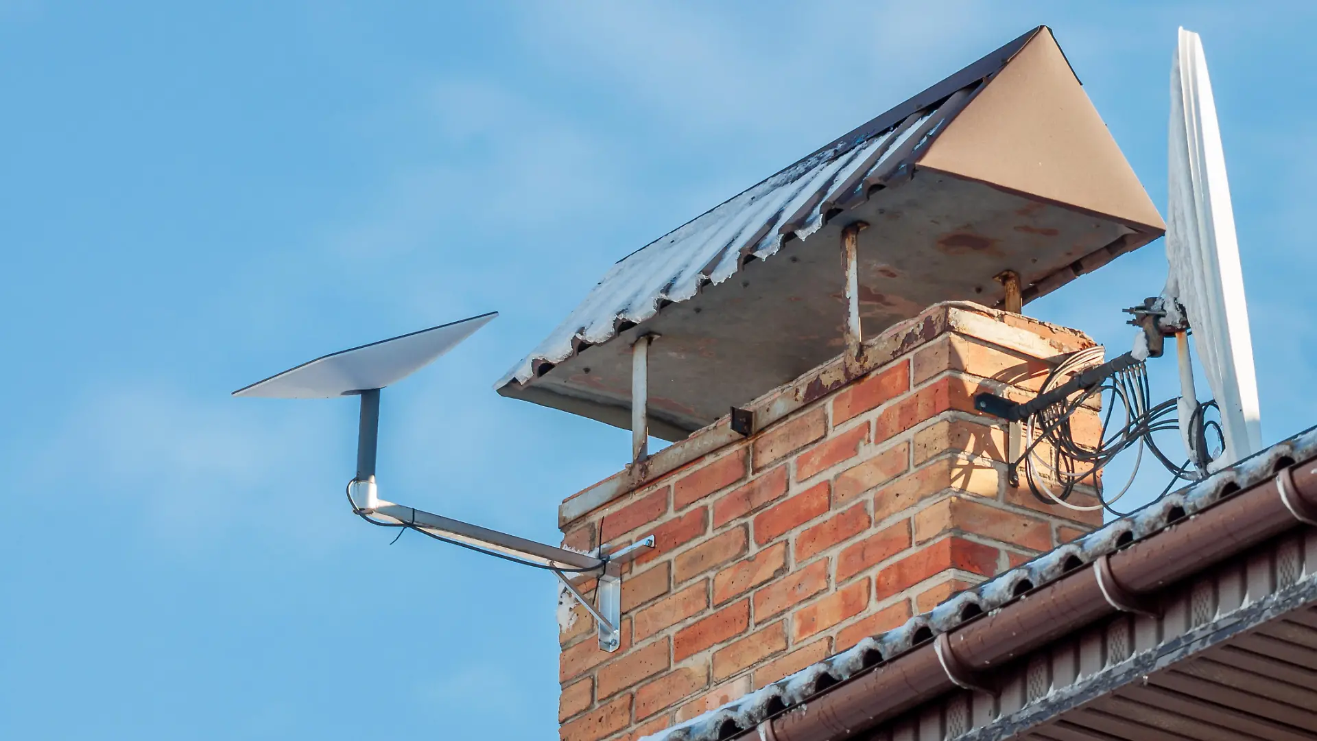 A residential roof featuring a brick chimney, old satellite dish, and a modern Starlink dish against a clear blue sky backdrop.