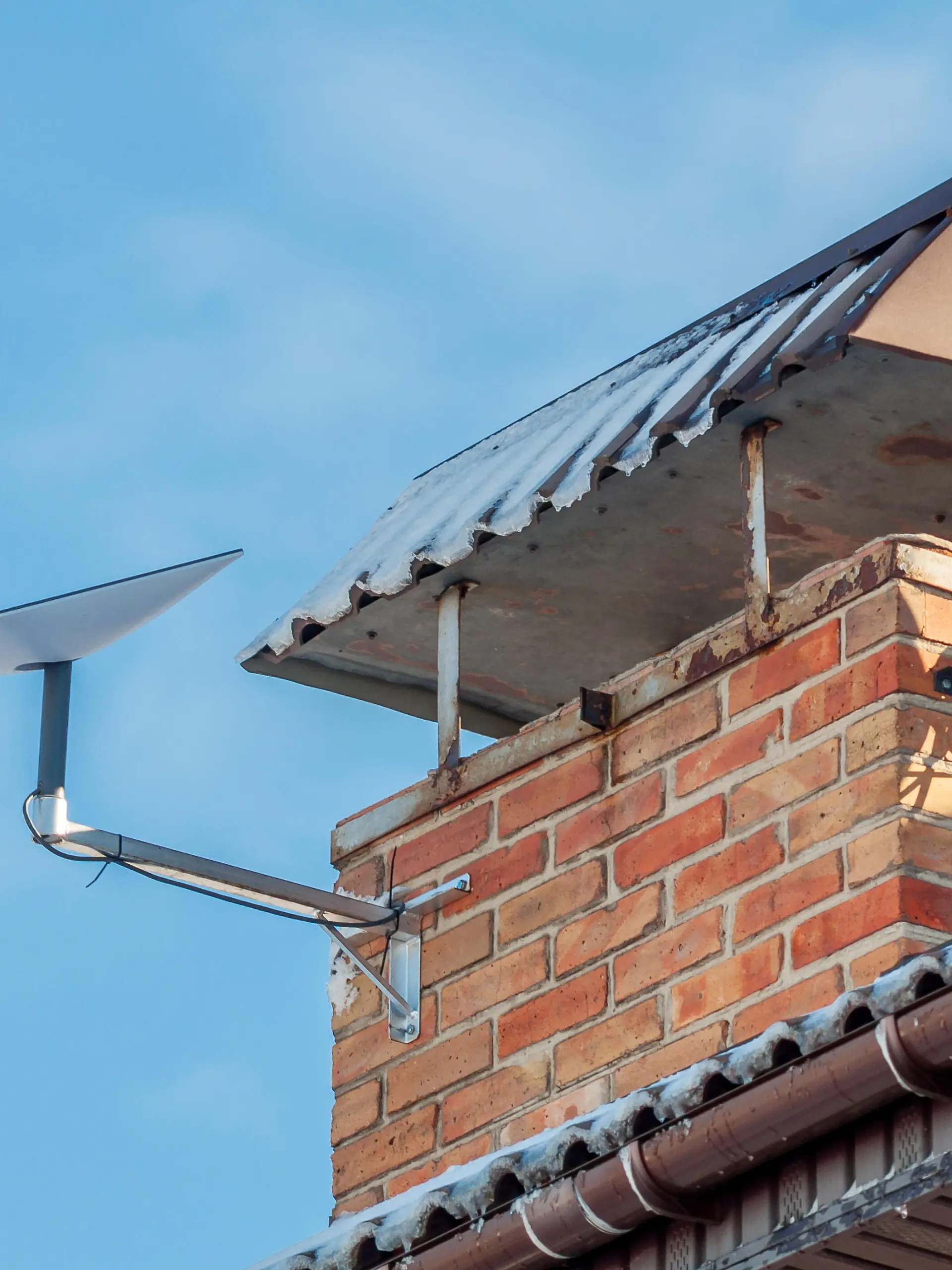 A residential roof featuring a brick chimney, old satellite dish, and a modern Starlink dish against a clear blue sky backdrop.