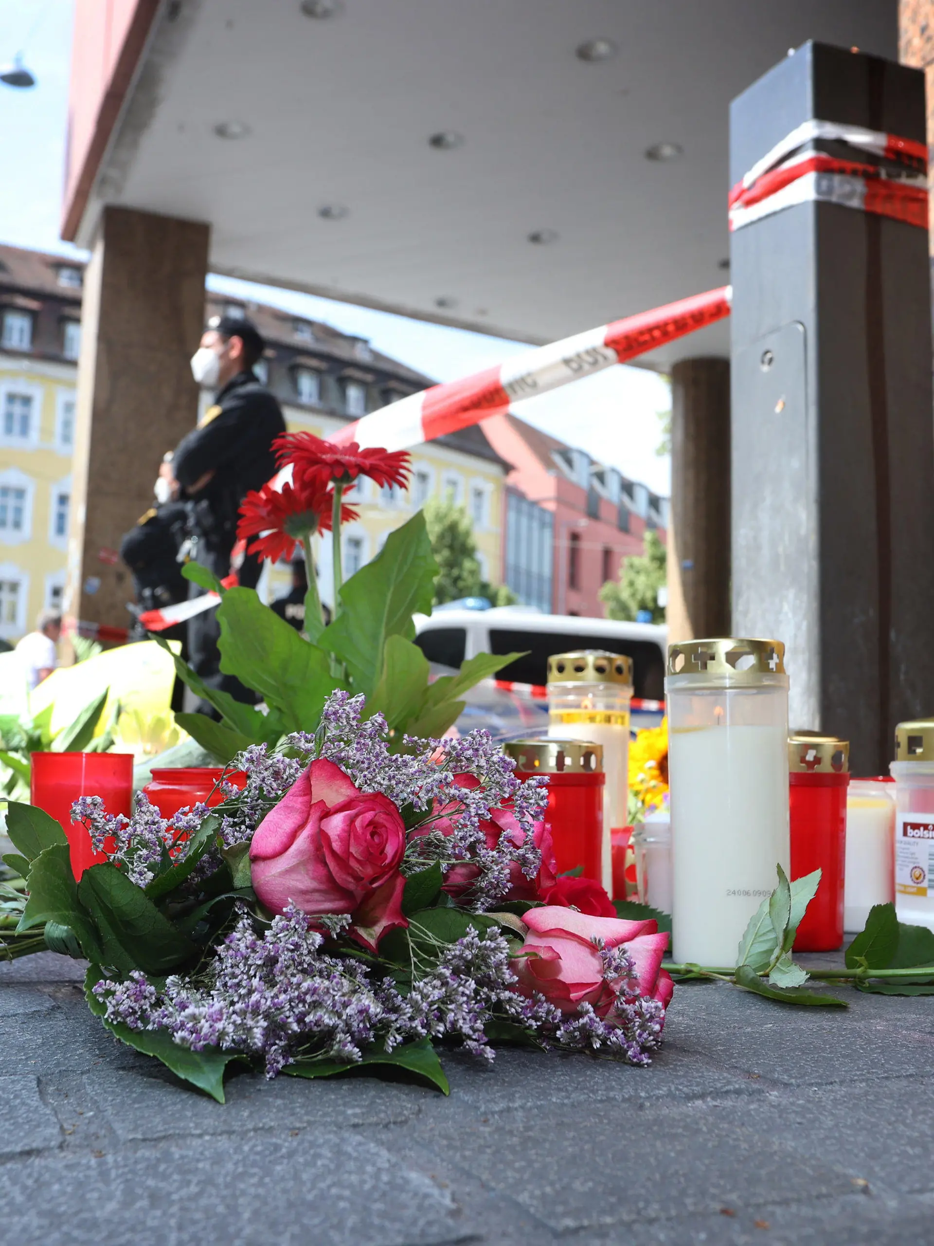 26.06.2021, Bayern, Würzburg: Ein Polizist steht neben Kerzen und Blumen vor einem geschlossenen und abgesperrten Geschäft in der Innenstadt. In Würzburg hat ein Mann am Vortag wahllos Menschen mit einem Messer attackiert. Drei Menschen starben, mindestens fünf wurden schwer verletzt. Foto: Karl-Josef Hildenbrand/dpa +++ dpa-Bildfunk +++