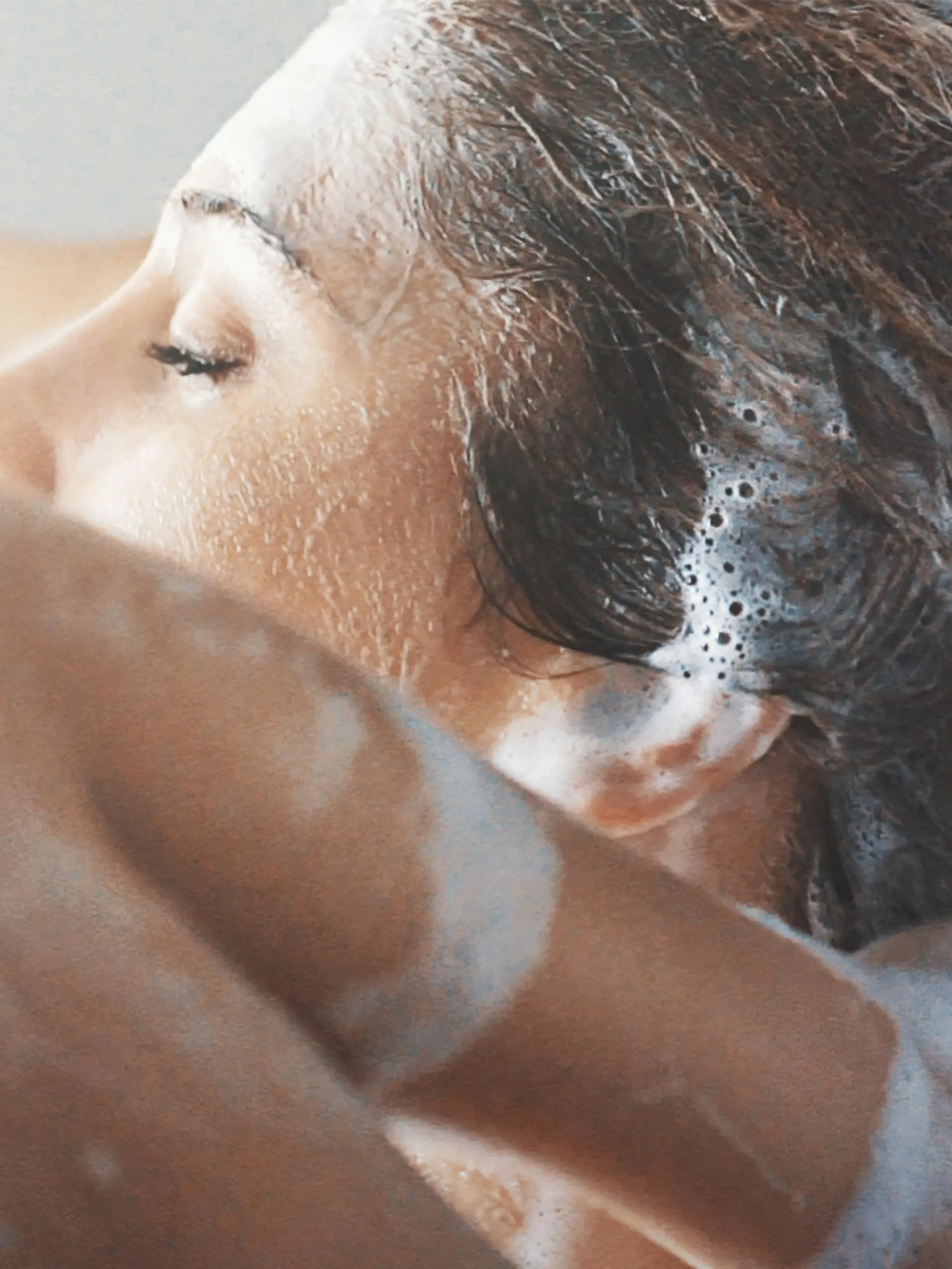 Closeup side view of mid 20's woman completely enjoying washiing her blond colored hair. She's going her finger through soapy hair, faced to the bathroom window.