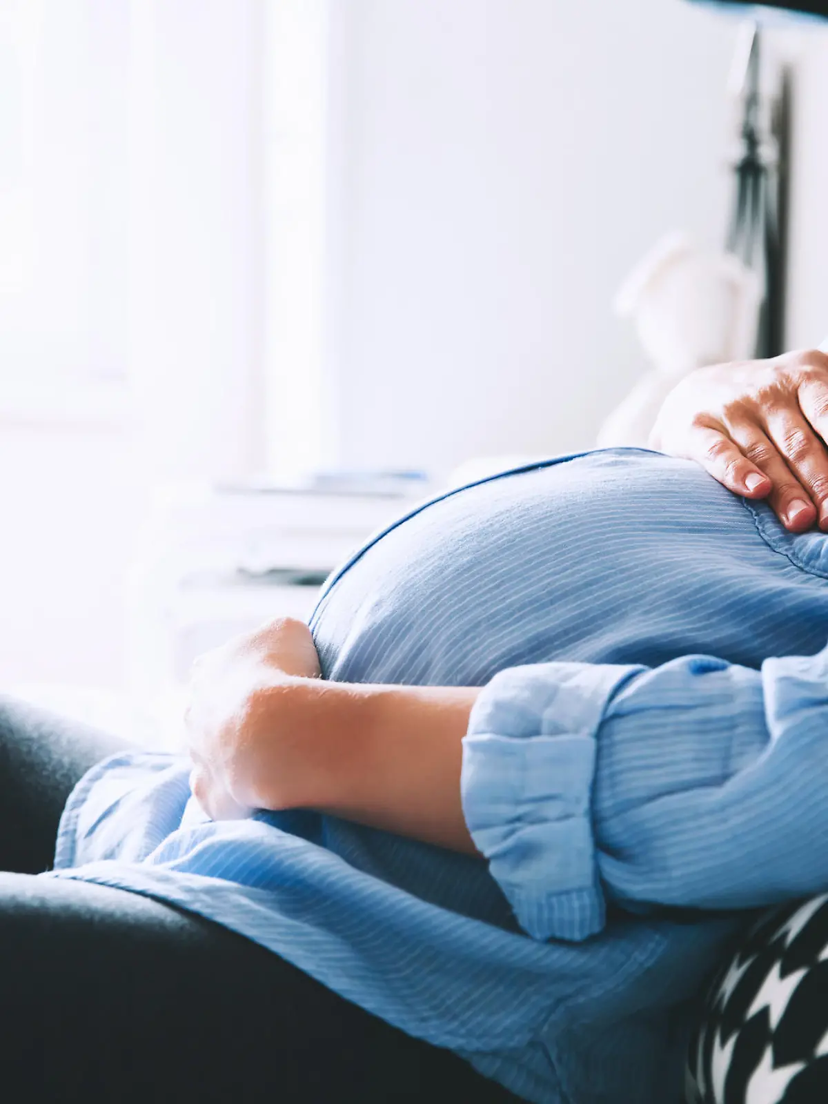 Beautiful pregnant woman sitting at bed and holds hands on belly in bedroom at home. Pregnancy, parenthood, preparation and expectation concept. Close-up, indoors.
