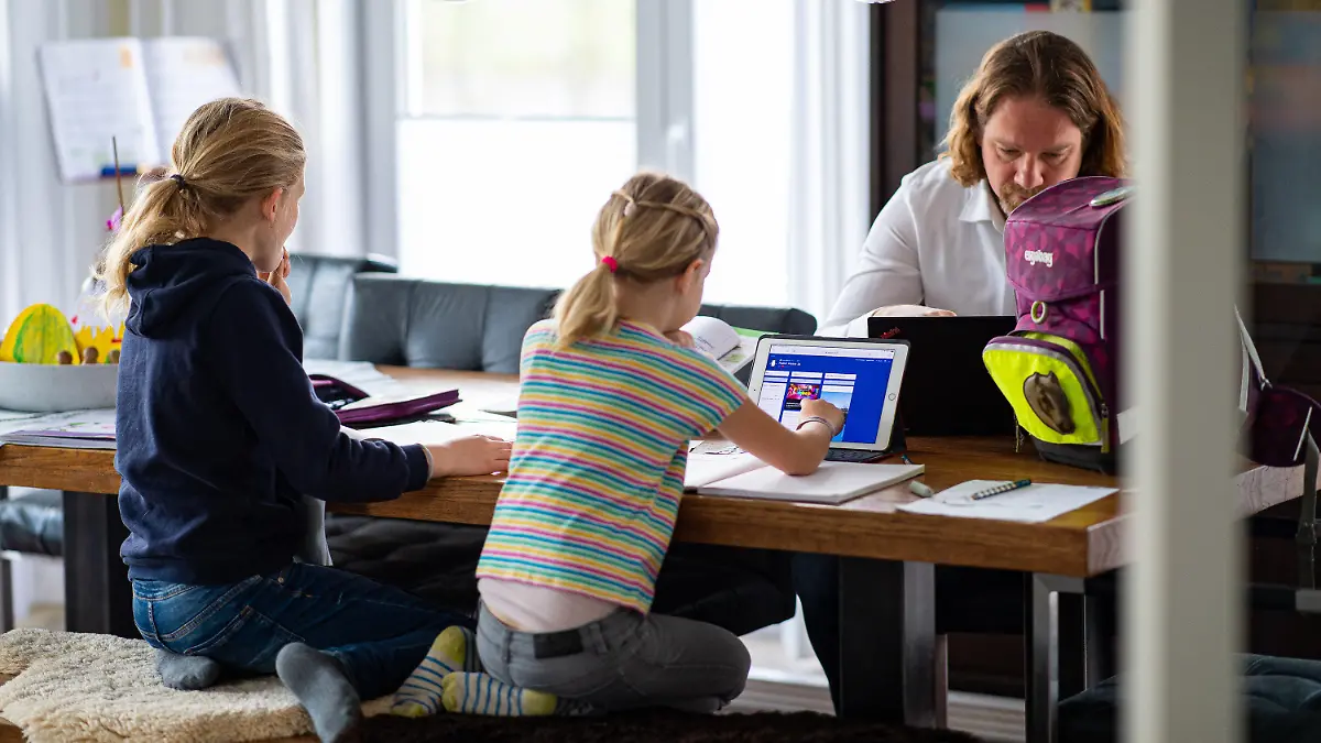 12.04.2021, Nordrhein-Westfalen, Warendorf: Die siebenjährige Karla (r) und ihre neunjährige Schwester Lotta sitzen im Esszimmer über ihren Hausaufgaben und werden von ihrem Vater Jens Bergmann unterstützt, während Mutter Stefanie (nicht auf dem Foto zu sehen) in der Küche am Computer im Homeoffice arbeitet. In Nordrhein-Westfalen sind die Osterferien zu Ende - ab Montag, 12.04.2021 haben die meisten Schüler Distanzunterricht, in Kitas gilt eingeschränkter Betrieb. Foto: Guido Kirchner/dpa - ACHTUNG: Nur zur einmaligen, redaktionellen Verwendung im Zusammenhang mit der aktuellen Berichterstattung über Homeschooling am 12.04.2021 und nur mit vollständiger Nennung des vorstehenden Credits; keine Archivierung +++ dpa-Bildfunk +++