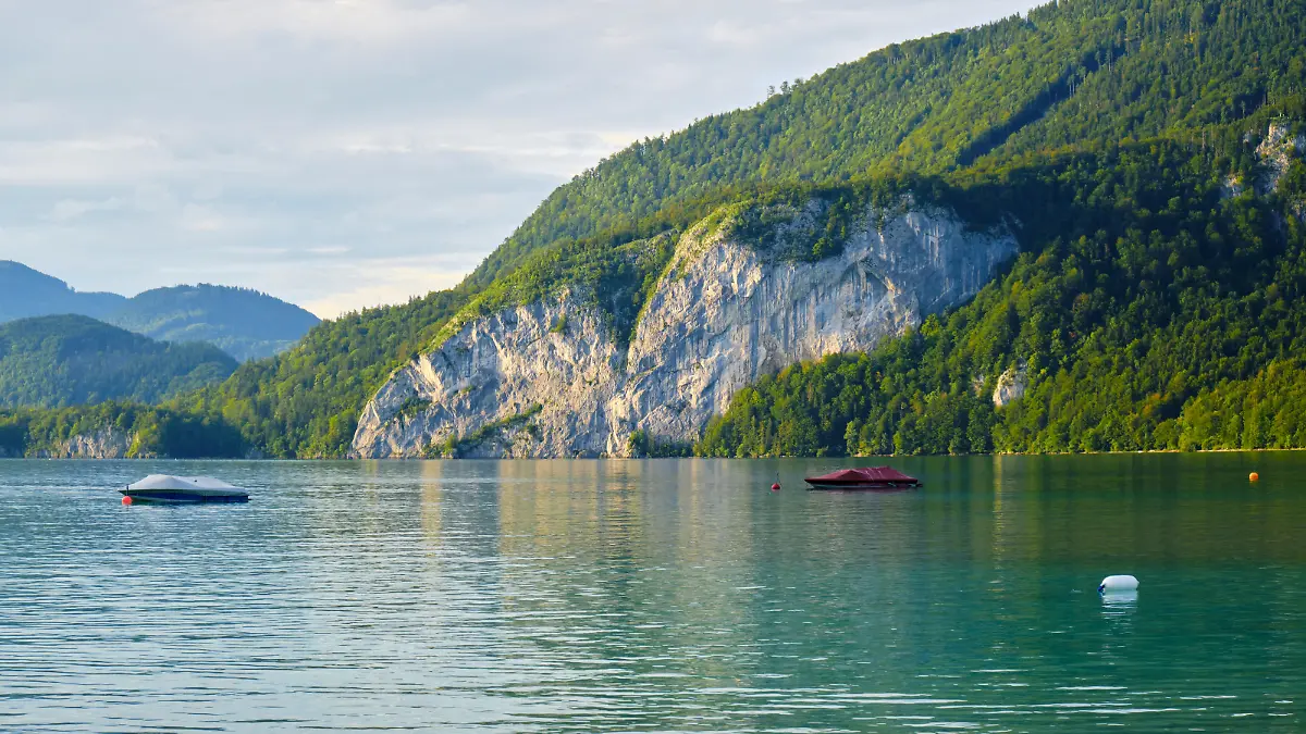 Calm waters of Wolfgang lake (Wolfgangsee) Austria, in the golden light of the sunset, with a couple of boats anchored - beautiful tall rock wall and forest in the background.