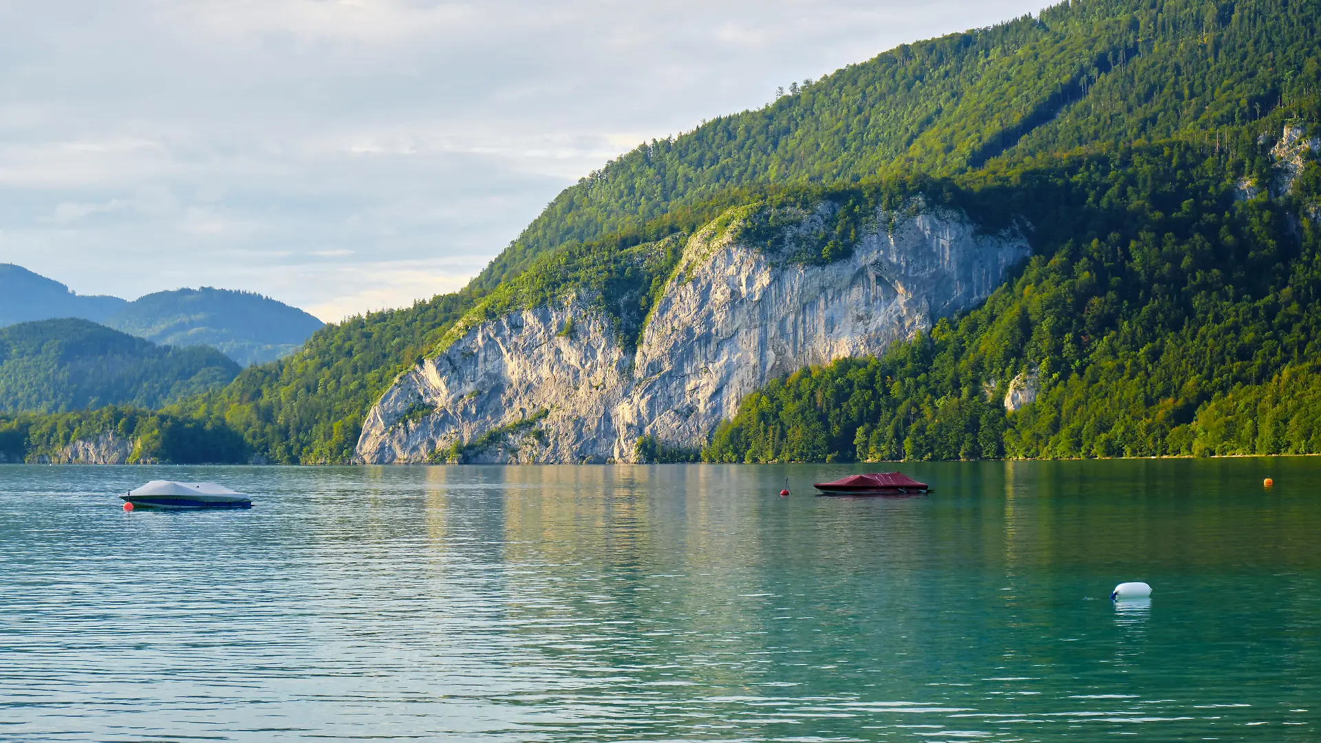 Calm waters of Wolfgang lake (Wolfgangsee) Austria, in the golden light of the sunset, with a couple of boats anchored - beautiful tall rock wall and forest in the background.