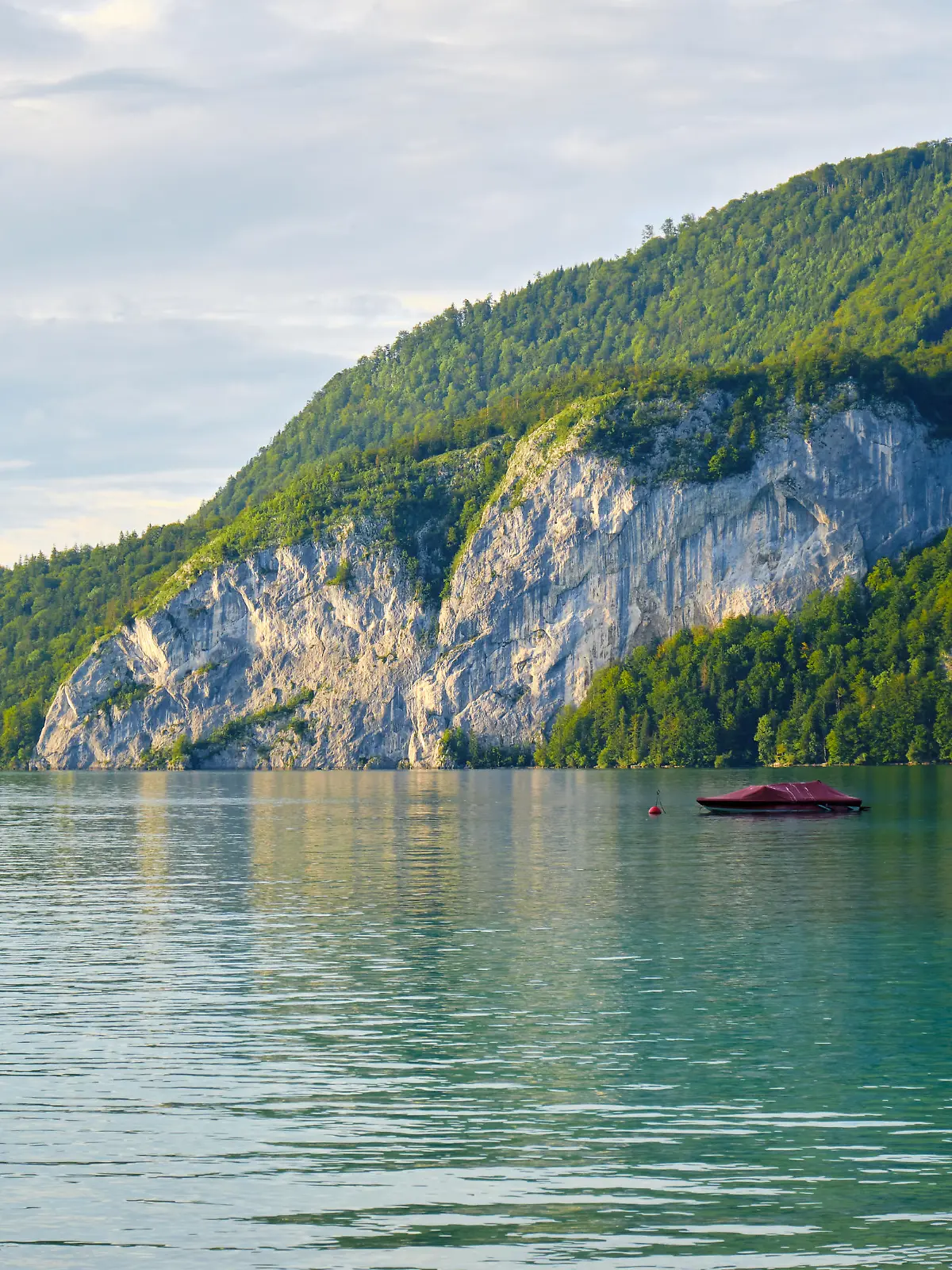 Calm waters of Wolfgang lake (Wolfgangsee) Austria, in the golden light of the sunset, with a couple of boats anchored - beautiful tall rock wall and forest in the background.