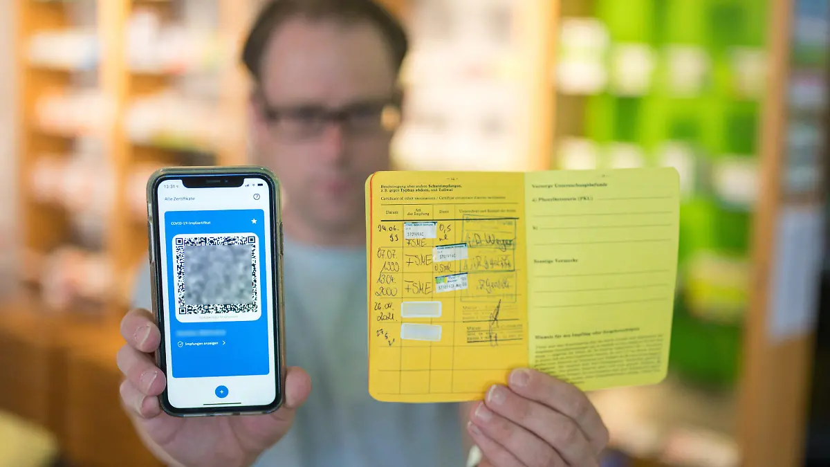 14.06.2021, Saarland, Saarbrücken: Apotheker Stefan Behrens hält ein Smartphone mit dem QR Code eines digitalen Impfausweises und ein Impfbuch in der Hand. Seit heute dürfen Apotheker den digitalen Impfausweis ausstellen. Foto: Oliver Dietze/dpa - ACHTUNG: Die Chargennummern des Coronaimpfstoffes wurden aus rechtlichen Gründen gepixelt +++ dpa-Bildfunk +++