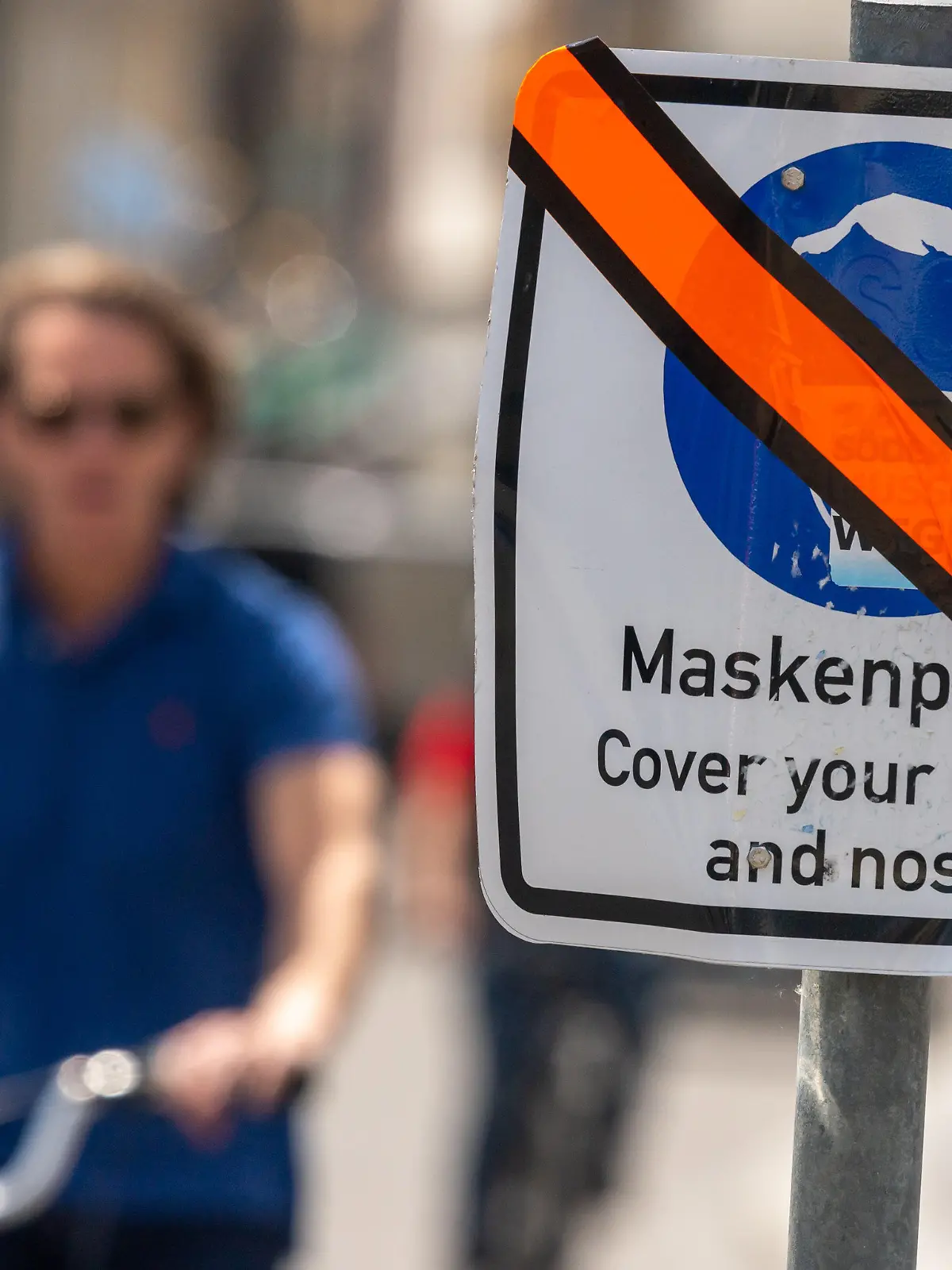 11.06.2021, Bayern, München: Ein mit orangen Klebeband durchgestrichenes Schild mit der Aufschrift «Maskenpflicht - Cover your mouth and nose!» steht in der Innenstadt am Straßenrand. Die generelle Maskenpflicht, in einem gekennzeichneten Bereich der Altstadt zwischen 9 und 21 Uhr galt, wurde aufgehoben. Foto: Peter Kneffel/dpa +++ dpa-Bildfunk +++