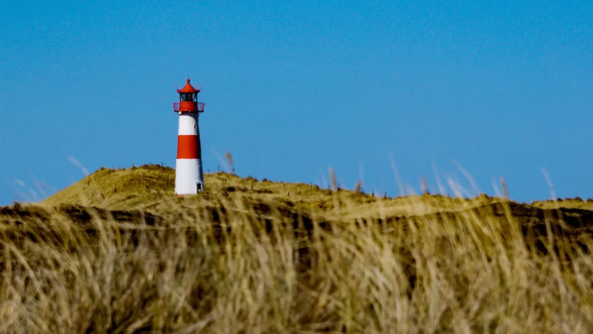 ARCHIV - 09.04.2019, Schleswig-Holstein, List: Die Sonne scheint am Strand von Sylt am sogenannten Ellenbogen auf einen Leuchtturm der zwischen den Dünen steht.  Am 1. Mai 2021 fährt Sylt als Teil der Modellregion Nordfriesland den Tourismus langsam und mit strengen Auflagen wieder hoch. Die Teilnahme ist auf der Insel umstritten und bringt erneut einen Konflikt an die Oberfläche, der schon lange auf der Insel gärte und im vergangenen Jahr nach dem Hochfahren des Tourismus von Null auf Hundert im Sommer hochkochte. (zu dpa: «Overtourism versus Nachhaltigkeit - Sylt sucht seinen Weg») Foto: Axel Heimken/dpa +++ dpa-Bildfunk +++