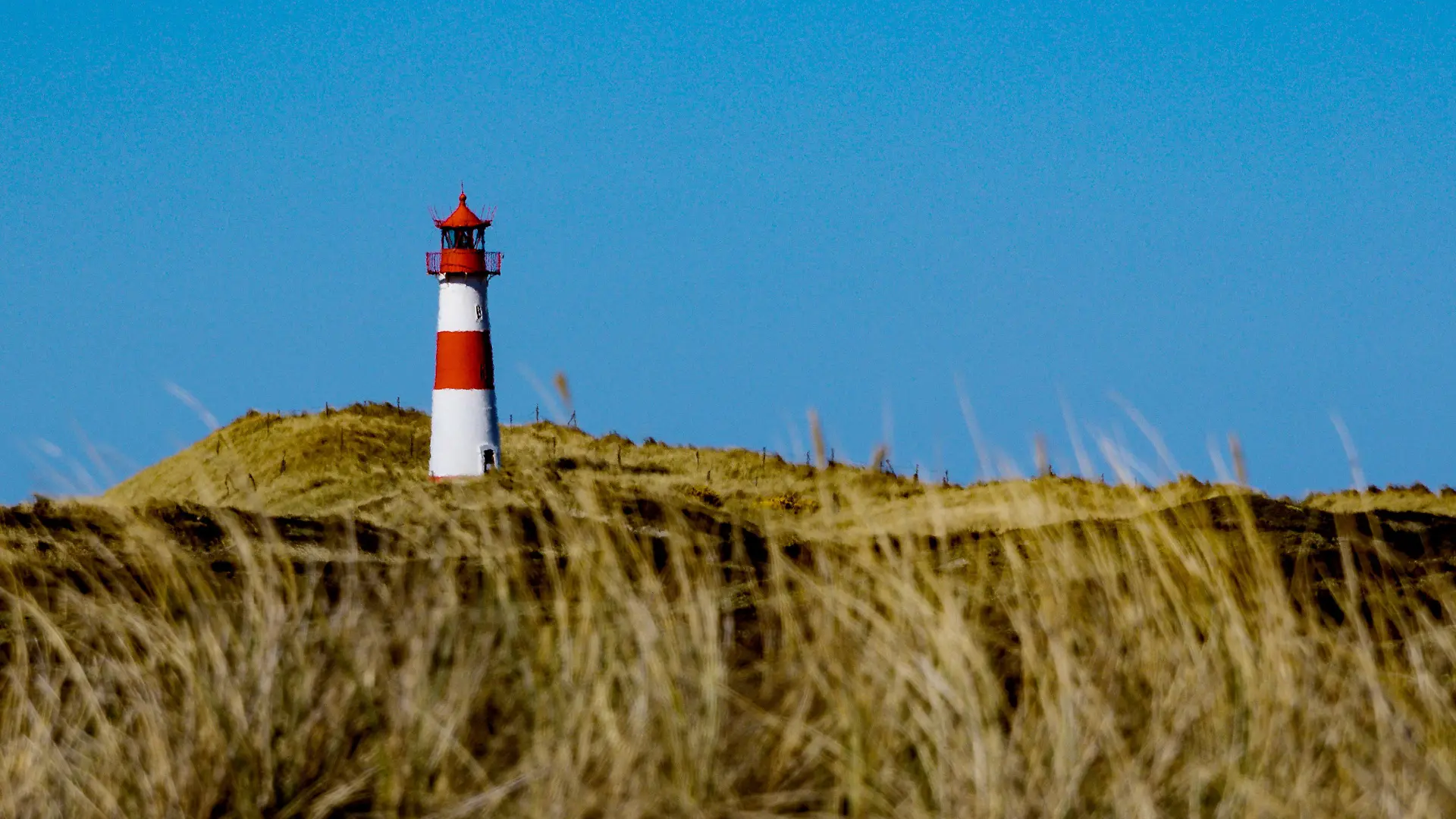 ARCHIV - 09.04.2019, Schleswig-Holstein, List: Die Sonne scheint am Strand von Sylt am sogenannten Ellenbogen auf einen Leuchtturm der zwischen den Dünen steht. Am 1. Mai 2021 fährt Sylt als Teil der Modellregion Nordfriesland den Tourismus langsam und mit strengen Auflagen wieder hoch. Die Teilnahme ist auf der Insel umstritten und bringt erneut einen Konflikt an die Oberfläche, der schon lange auf der Insel gärte und im vergangenen Jahr nach dem Hochfahren des Tourismus von Null auf Hundert im Sommer hochkochte. (zu dpa: «Overtourism versus Nachhaltigkeit - Sylt sucht seinen Weg») Foto: Axel Heimken/dpa +++ dpa-Bildfunk +++