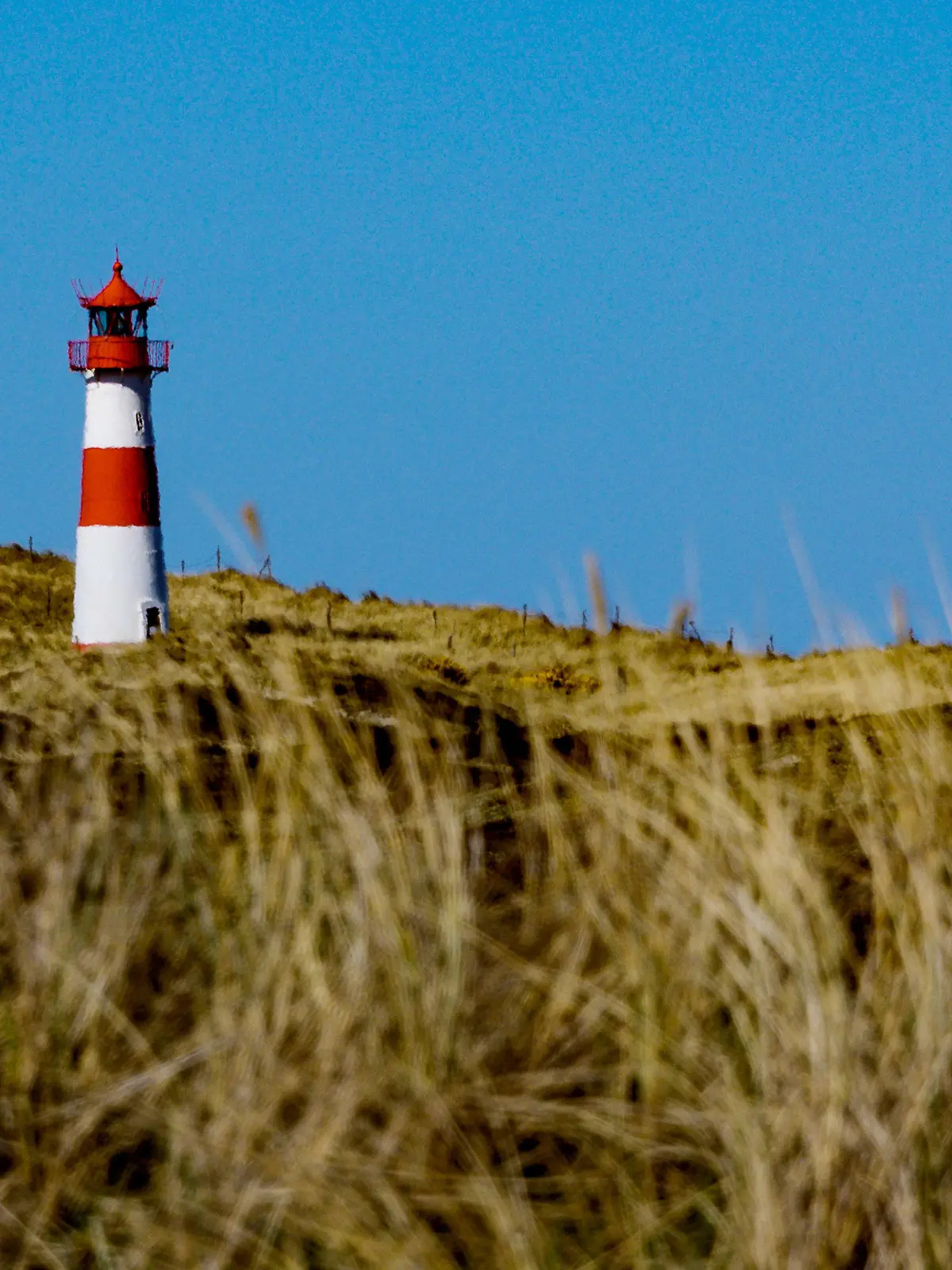 ARCHIV - 09.04.2019, Schleswig-Holstein, List: Die Sonne scheint am Strand von Sylt am sogenannten Ellenbogen auf einen Leuchtturm der zwischen den Dünen steht.  Am 1. Mai 2021 fährt Sylt als Teil der Modellregion Nordfriesland den Tourismus langsam und mit strengen Auflagen wieder hoch. Die Teilnahme ist auf der Insel umstritten und bringt erneut einen Konflikt an die Oberfläche, der schon lange auf der Insel gärte und im vergangenen Jahr nach dem Hochfahren des Tourismus von Null auf Hundert im Sommer hochkochte. (zu dpa: «Overtourism versus Nachhaltigkeit - Sylt sucht seinen Weg») Foto: Axel Heimken/dpa +++ dpa-Bildfunk +++